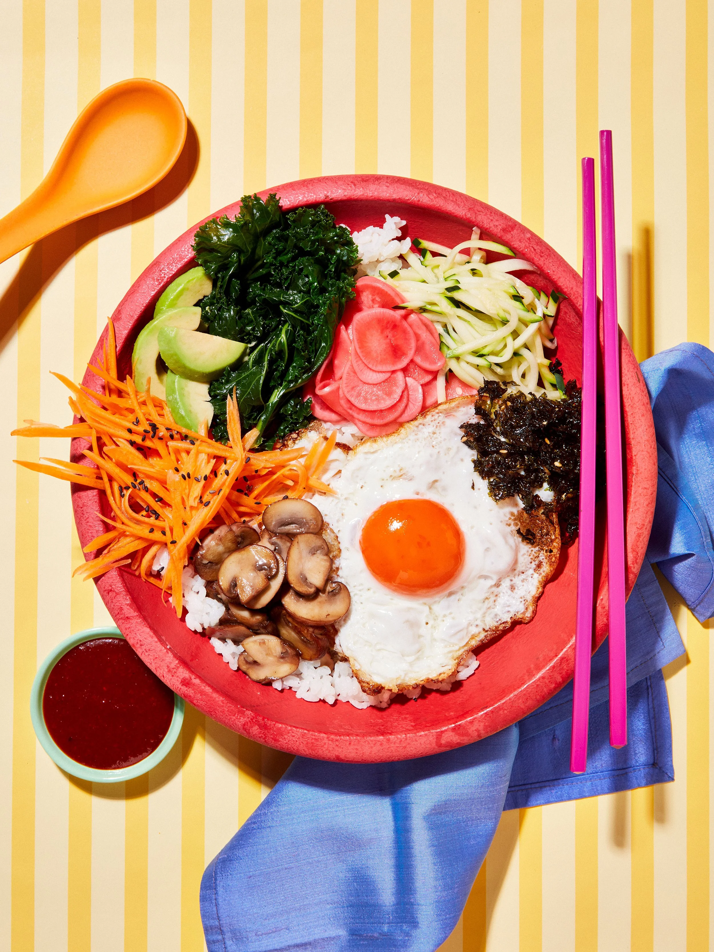 Colorful bibimbap bowl with rice, fried egg, avocado, kale, carrots, mushrooms, radishes, zucchini, and seaweed, served with chopsticks and sauce