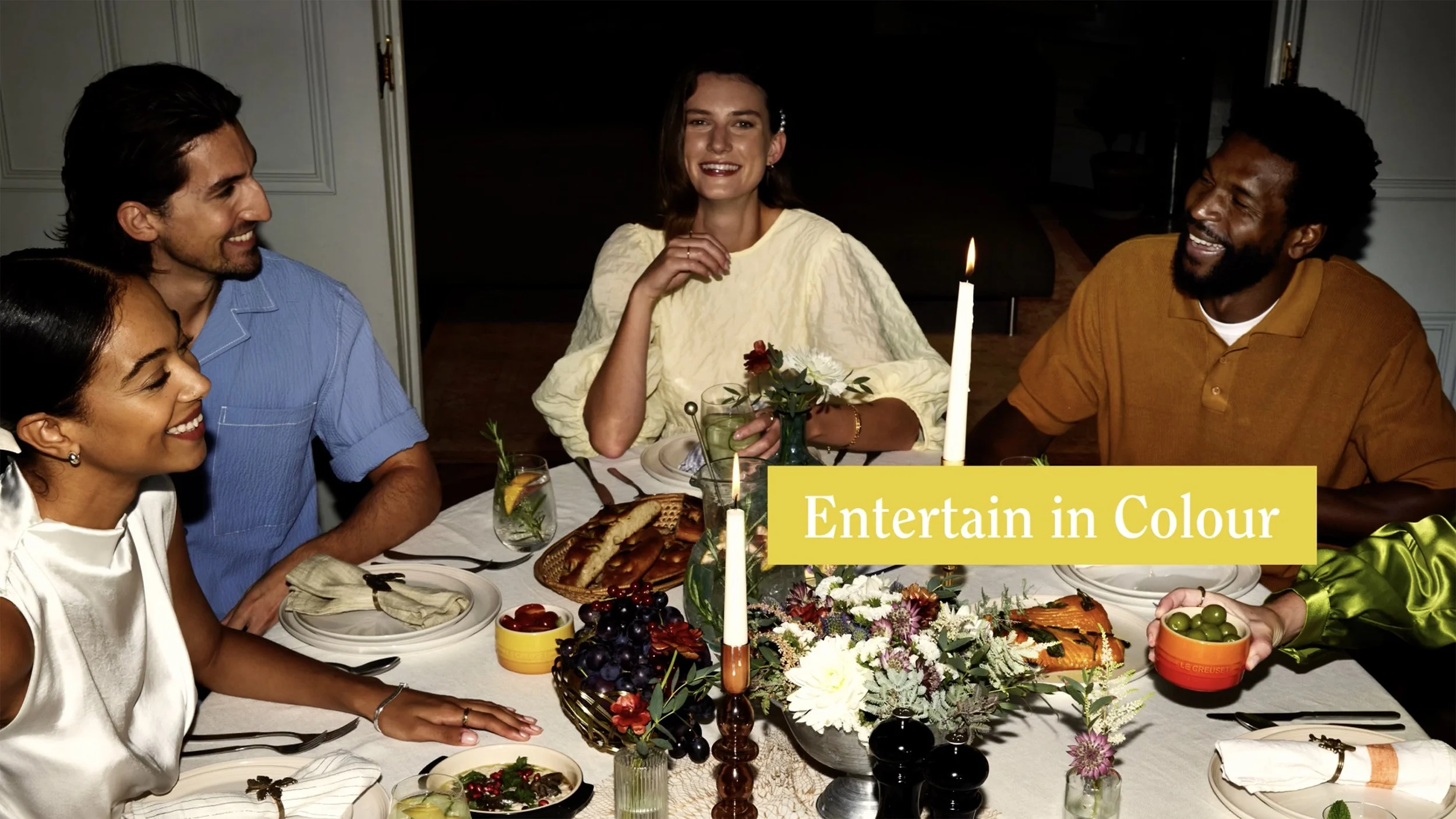 Friends laughing around a colorful dinner table with flowers, candles, food, and drinks; yellow text reads “Entertain in Colour.”