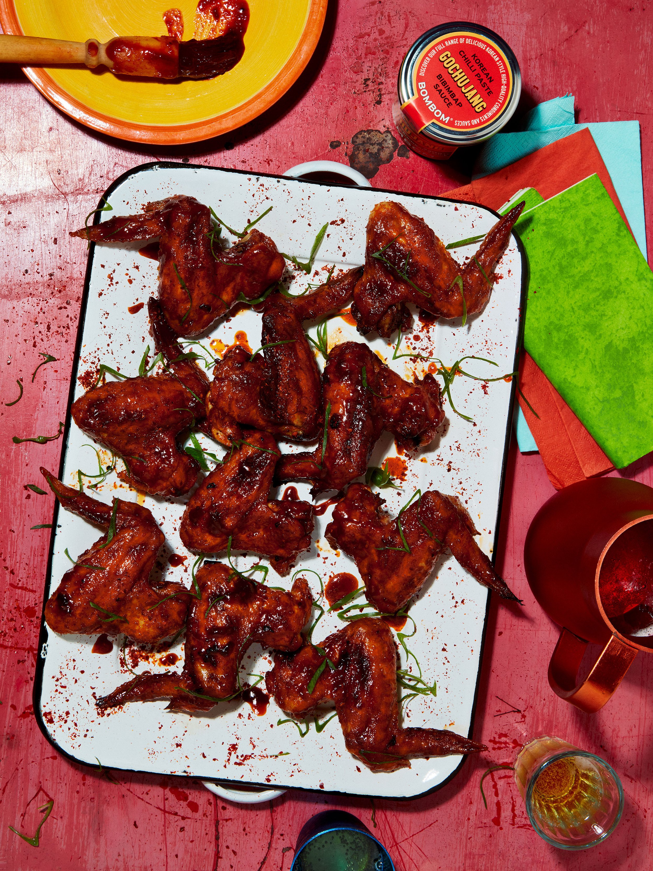 Korean gochujang-glazed chicken wings garnished with scallions on a white platter, with a tub of gochujang sauce and colorful napkins.
