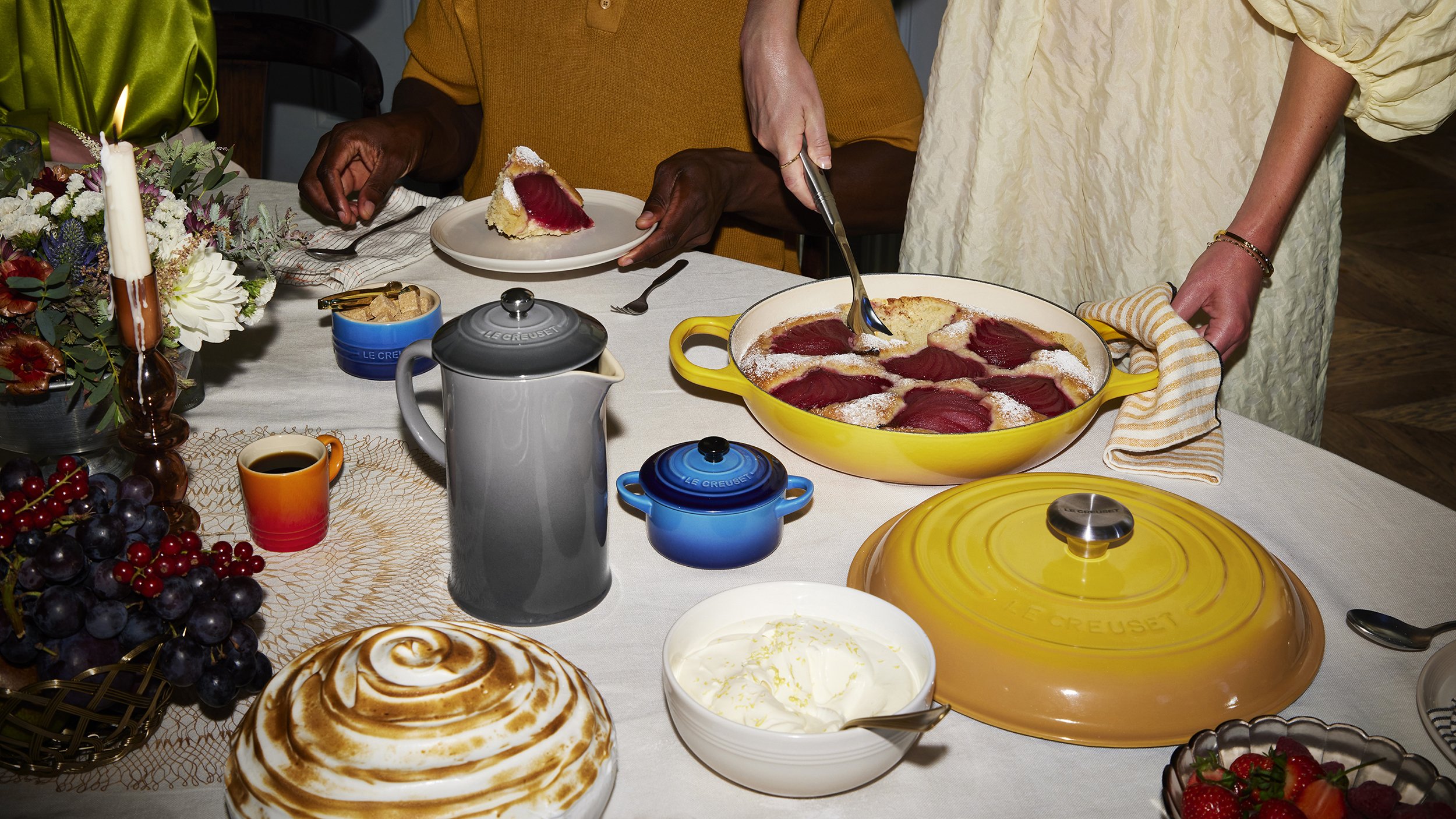 Festive dessert table with a berry tart, sliced cake, coffee, fruit, flowers, and colorful cookware.