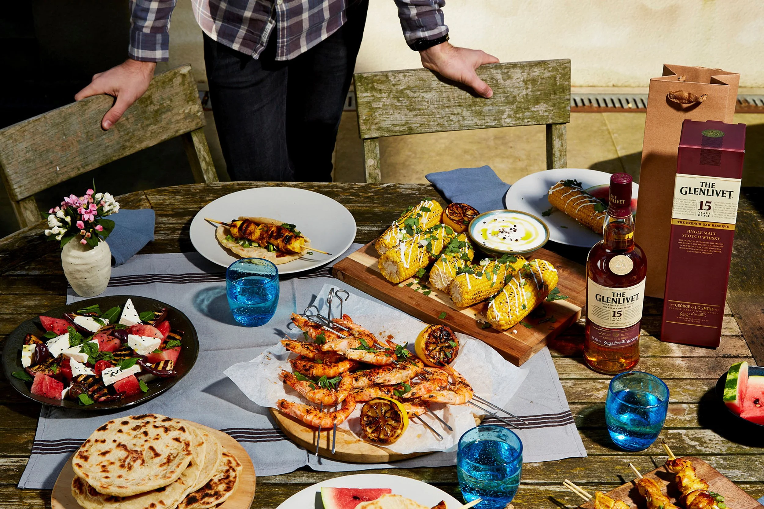 Outdoor barbecue spread with grilled shrimp skewers, corn, flatbreads, salads, dipping sauce, and Glenlivet 15-year Scotch whisky