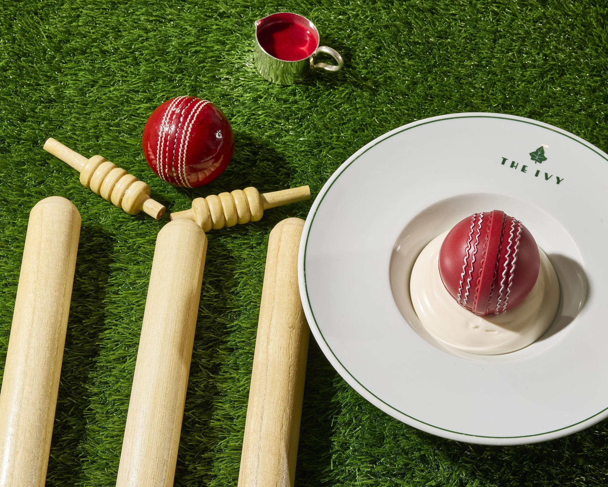 Cricket-themed still life with red cricket balls, wooden cricket bats, a silver cup of red liquid, and a plated red cricket-ball dessert on artificial grass.