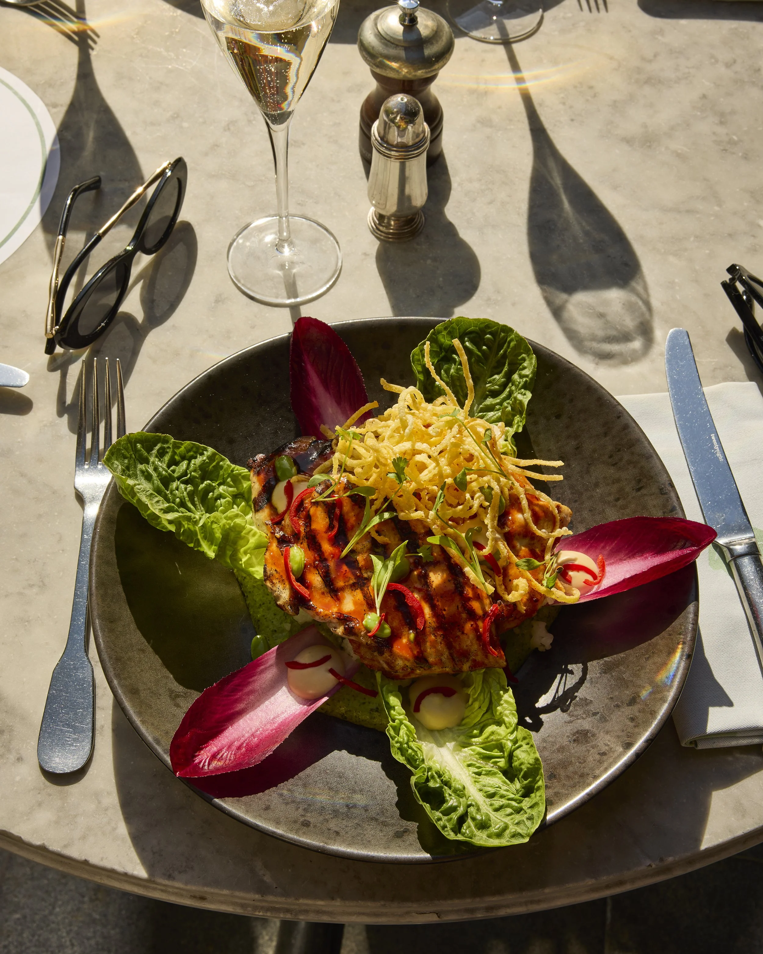 Grilled chicken dish with crispy fried onions, lettuce, peppers, and sauce on a black plate beside a glass of wine and table settings