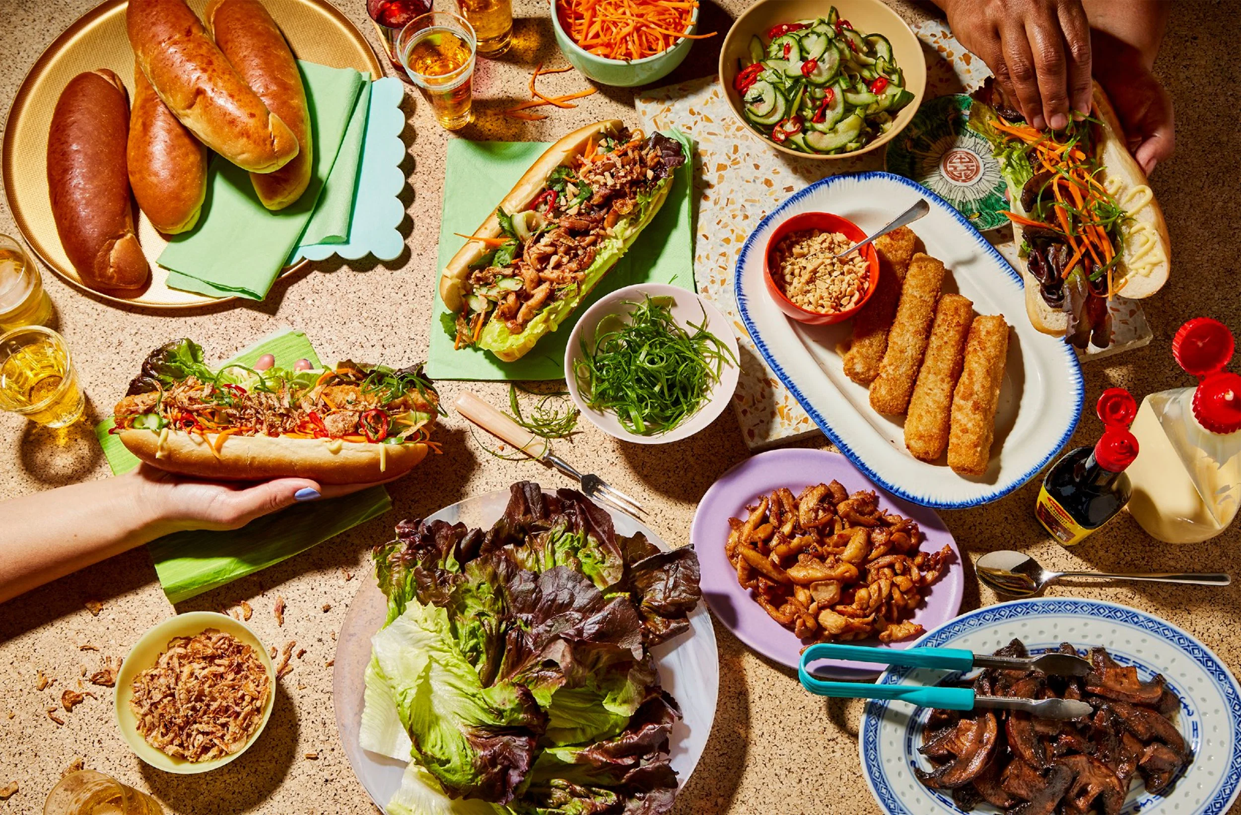 Overhead spread of Vietnamese food including banh mi sandwiches, spring rolls, grilled meats, fried chicken, fresh lettuce, pickled vegetables, herbs, and dipping sauces.