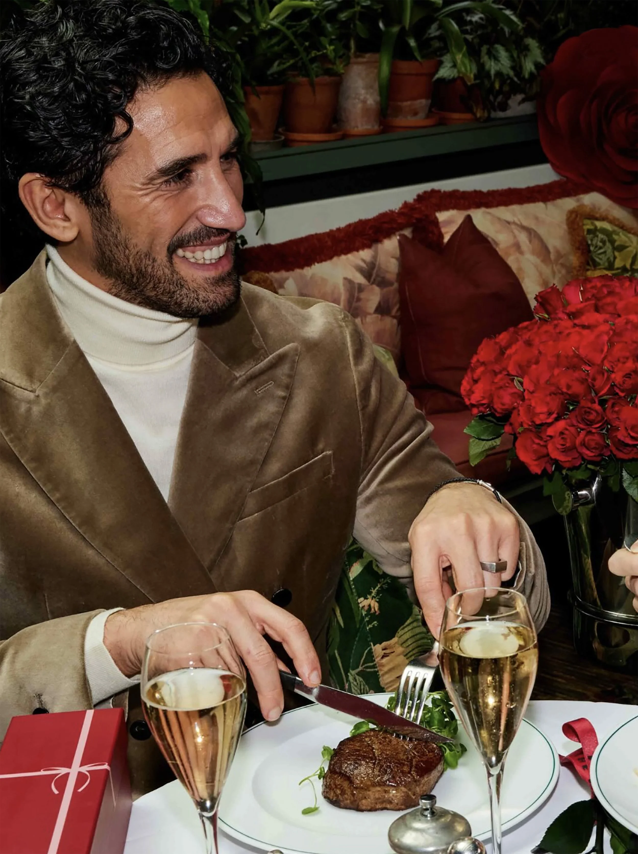 Smiling man in a brown jacket dining with champagne, steak, and red roses at a restaurant