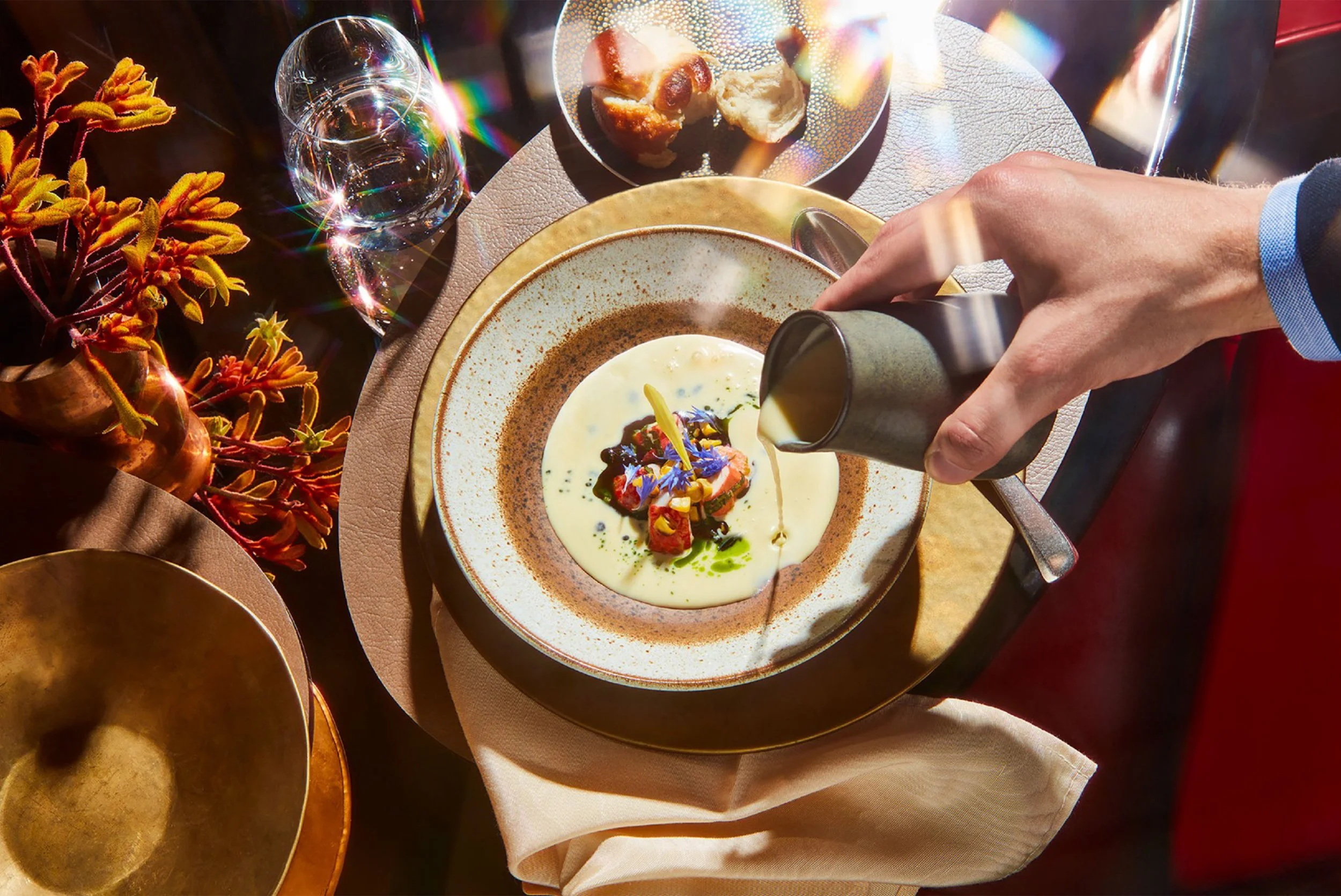 Fine dining table with a plated gourmet dish, creamy sauce being poured, wine glasses, bread, and flowers in warm sunlight