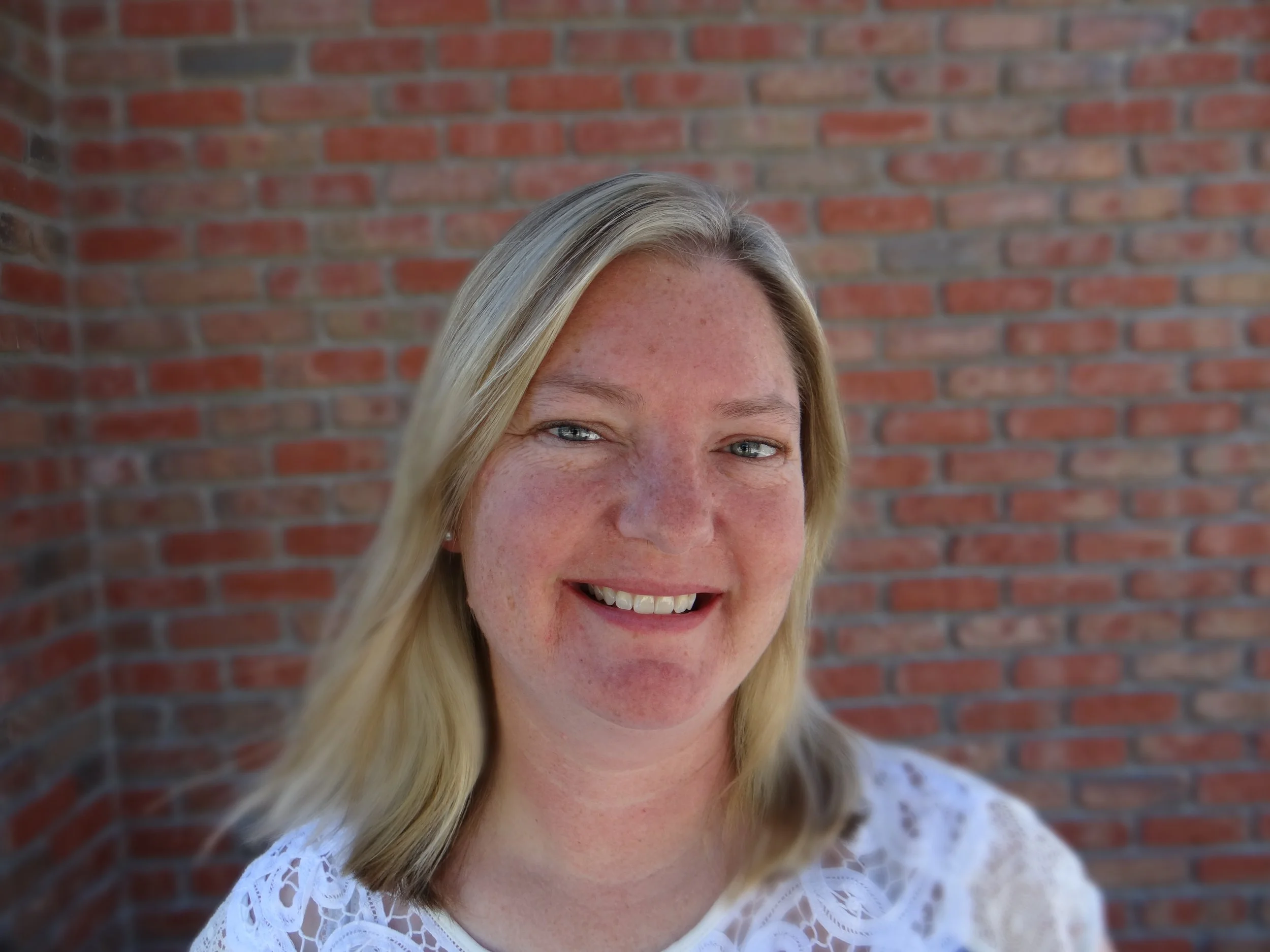 Smiling blonde woman in a white lace top posing in front of a brick wall