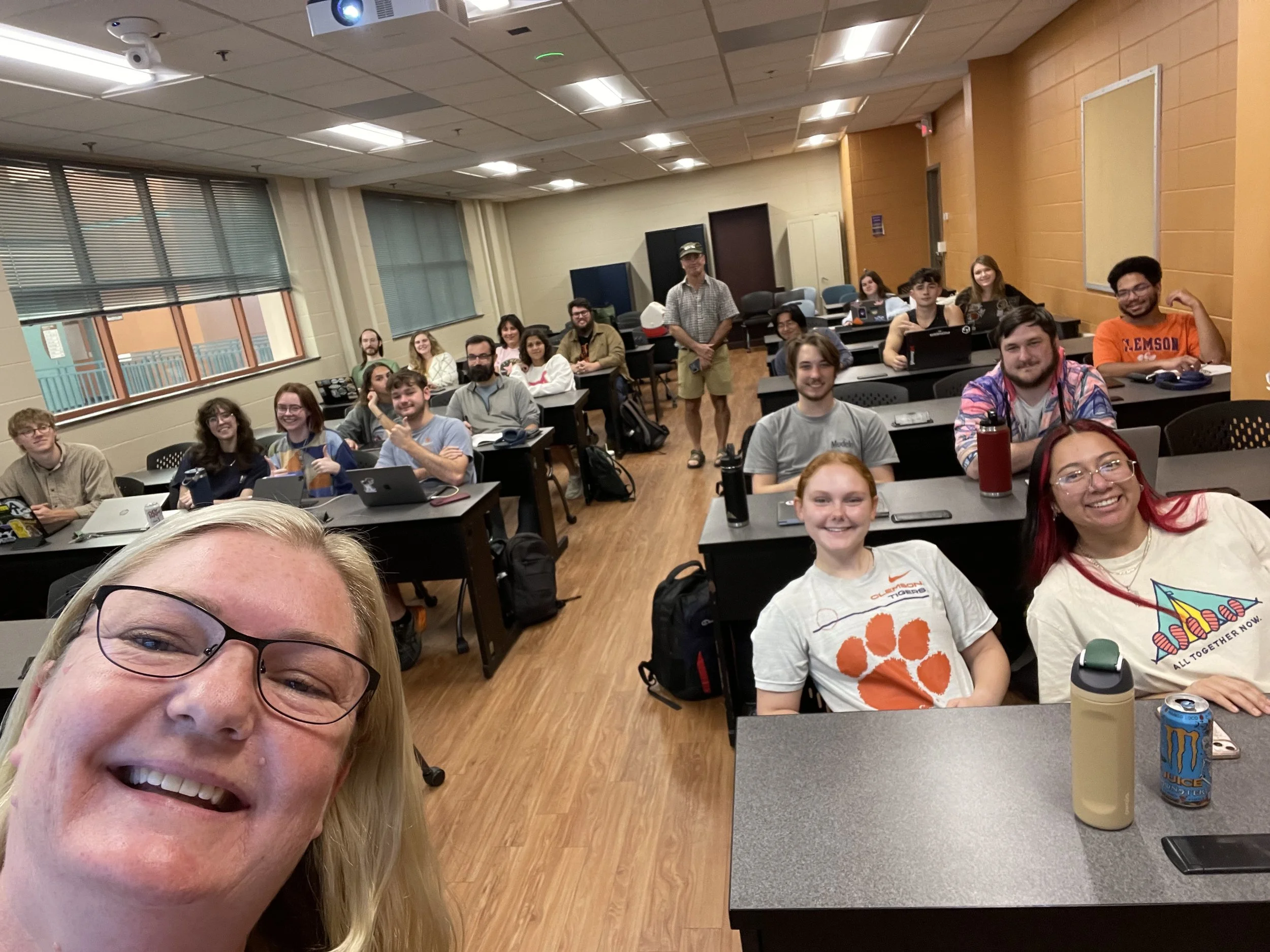 Group selfie of college students and an instructor in a classroom with desks, laptops, and a ceiling-mounted projector