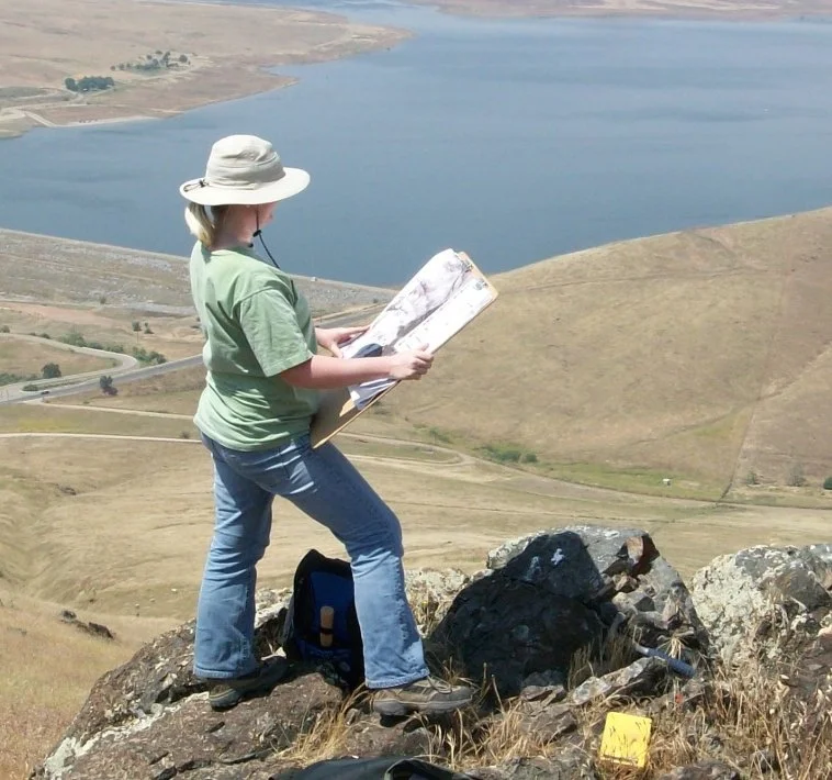 Hiker wearing a sun hat studies a map on rocky terrain overlooking a large reservoir and dry hills.