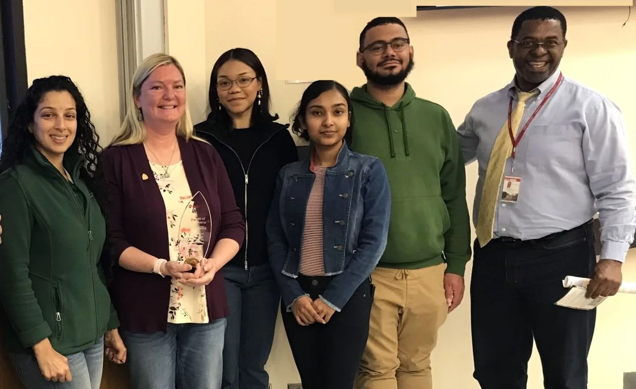 Group of six diverse colleagues posing indoors, with one woman holding a glass award.
