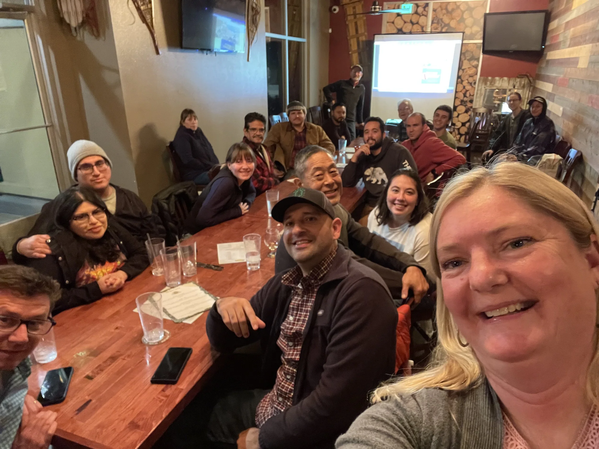 Group selfie of friends gathered around a restaurant table during a presentation, with a projector screen and fireplace in the background.