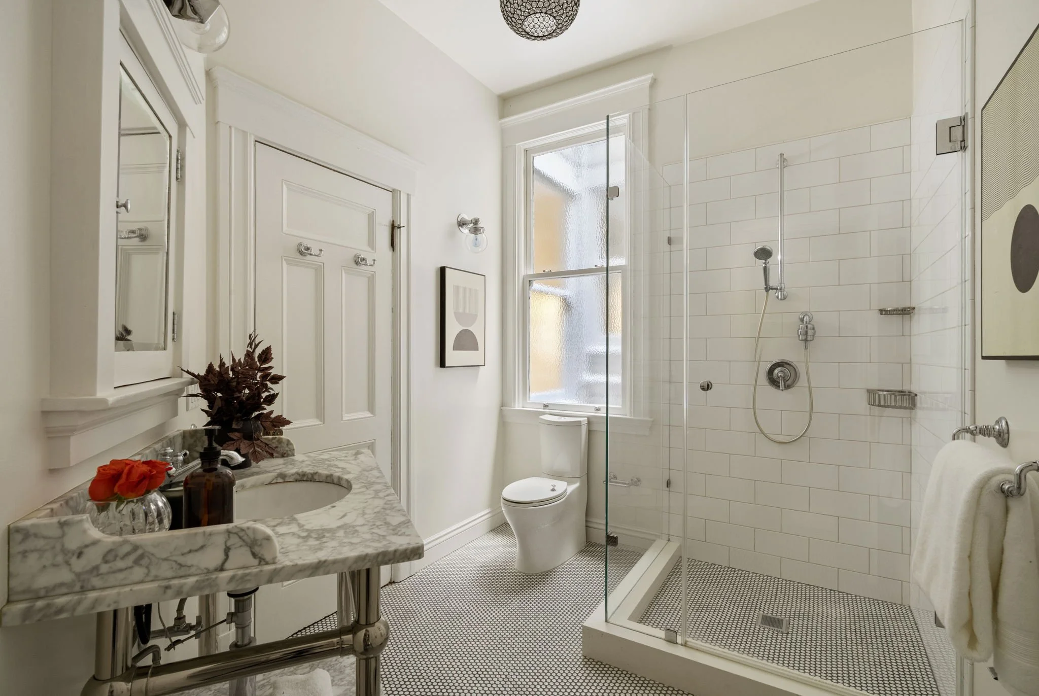 A white bathroom with a marble countertop sink, a large mirror, a small plant, and a soap dispenser. Next to it is a white toilet, a window, and a glass-enclosed shower area with white tiled walls and a handheld showerhead. There is a wall art piece 