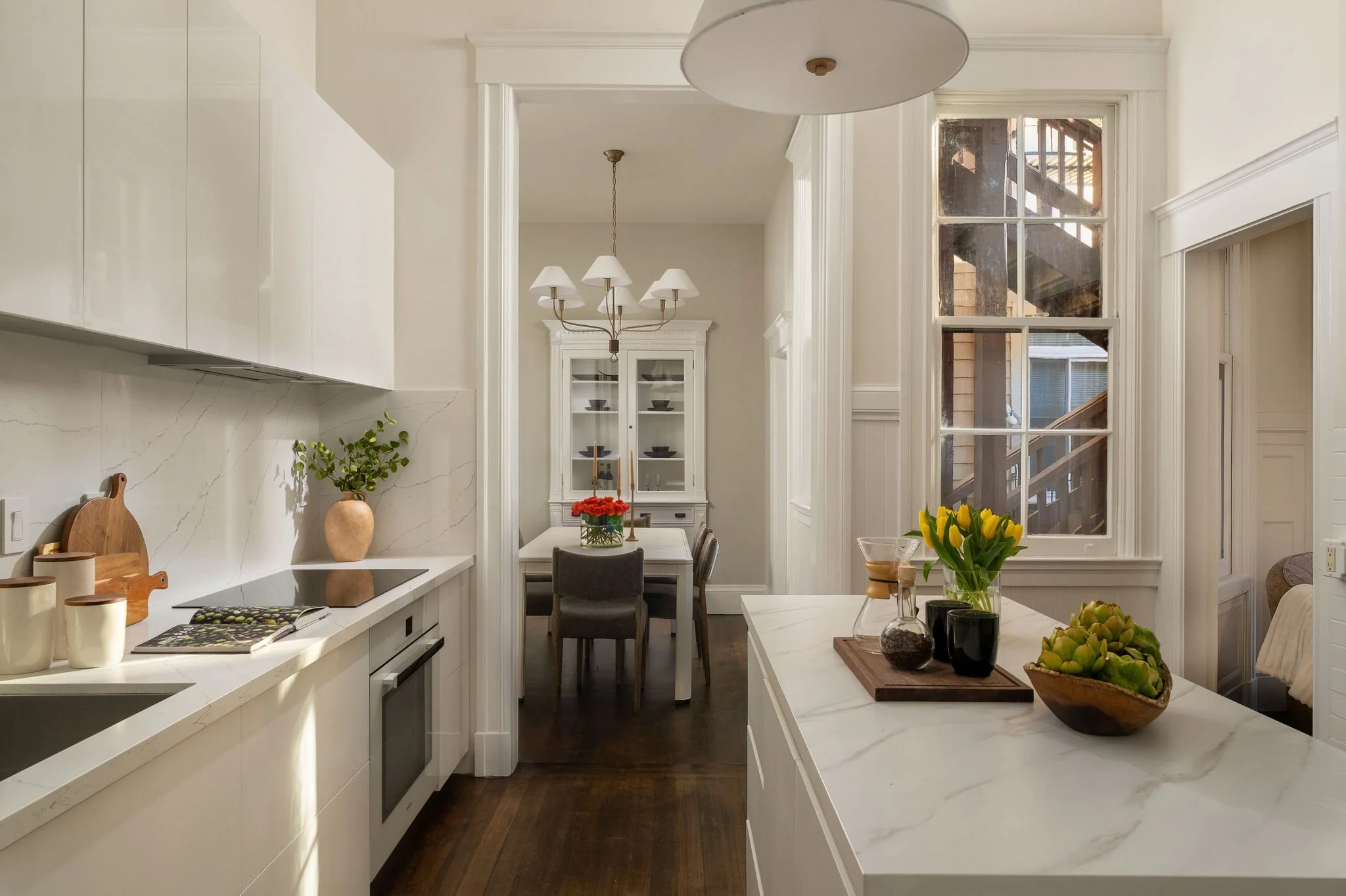 A bright, modern kitchen and dining area with white cabinetry, marble countertops, a large window, and decorative plants and flowers.