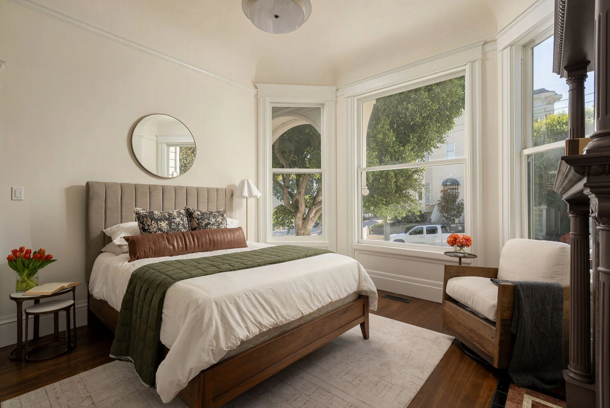 A cozy bedroom with a large bed, beige headboard, and white bedding with a green throw. Side table with a vase of red tulips and open book. Large corner window with white trim showing trees and houses outside. Circular mirror on wall, white lamp, woo