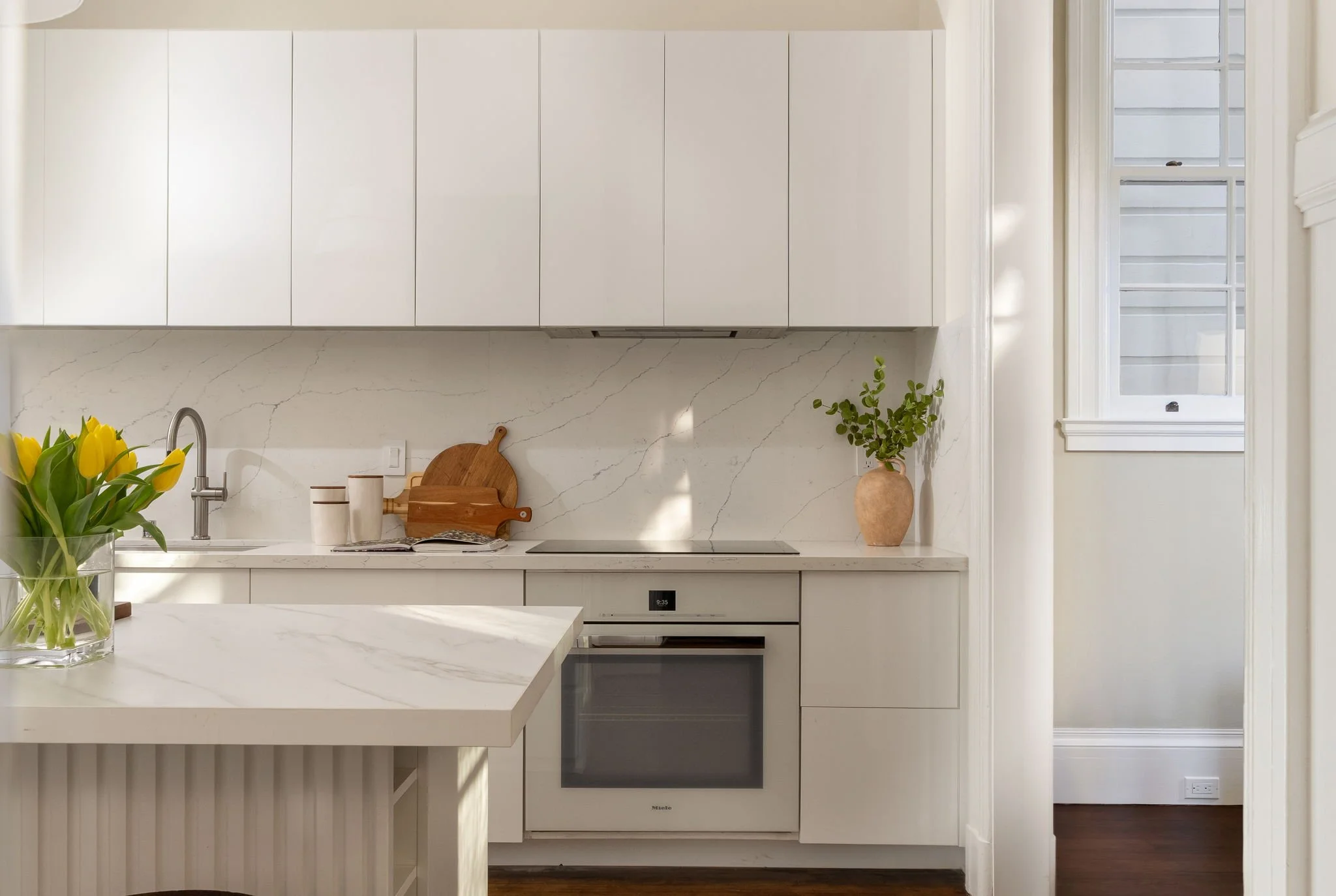 Modern kitchen featuring white cabinetry with minimalist design, quartz countertops, a marble backsplash, a built-in oven, and a small potted plant near a window.