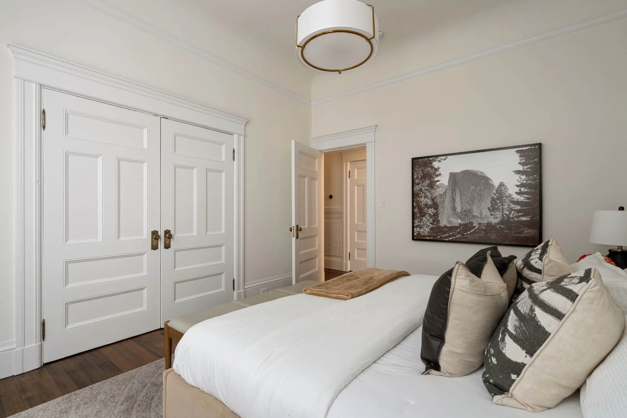 A bedroom with a white bed, various decorative pillows, framed landscape photo, white double closet doors, a door leading to another room, ceiling light fixture, dark wooden floor, and neutral wall color.