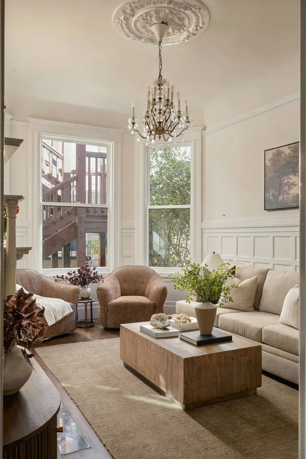 Living room with large windows, beige sofa, pink armchairs, a wooden coffee table, a chandelier, and decorating plants.
