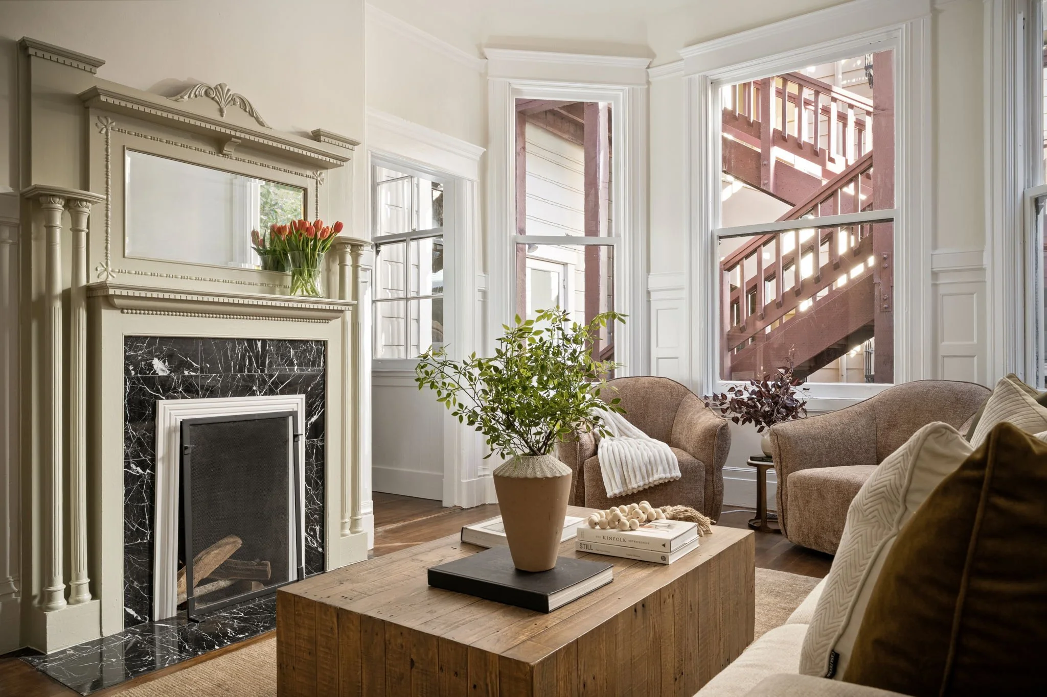 A cozy living room with a fireplace, beige armchairs, potted plants, and a coffee table with books, large windows revealing a staircase outside, and decorative crown molding.
