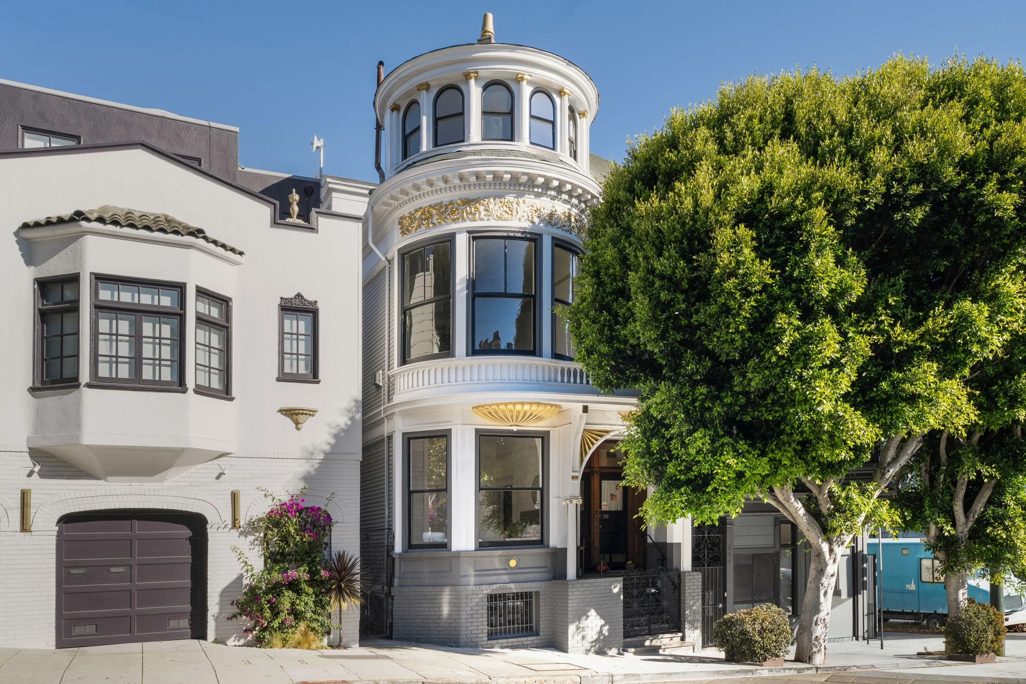 A white multi-story house with ornate architectural details, large rounded bay windows, a decorative balcony, and a prominent turret, partially obscured by a large leafy tree.