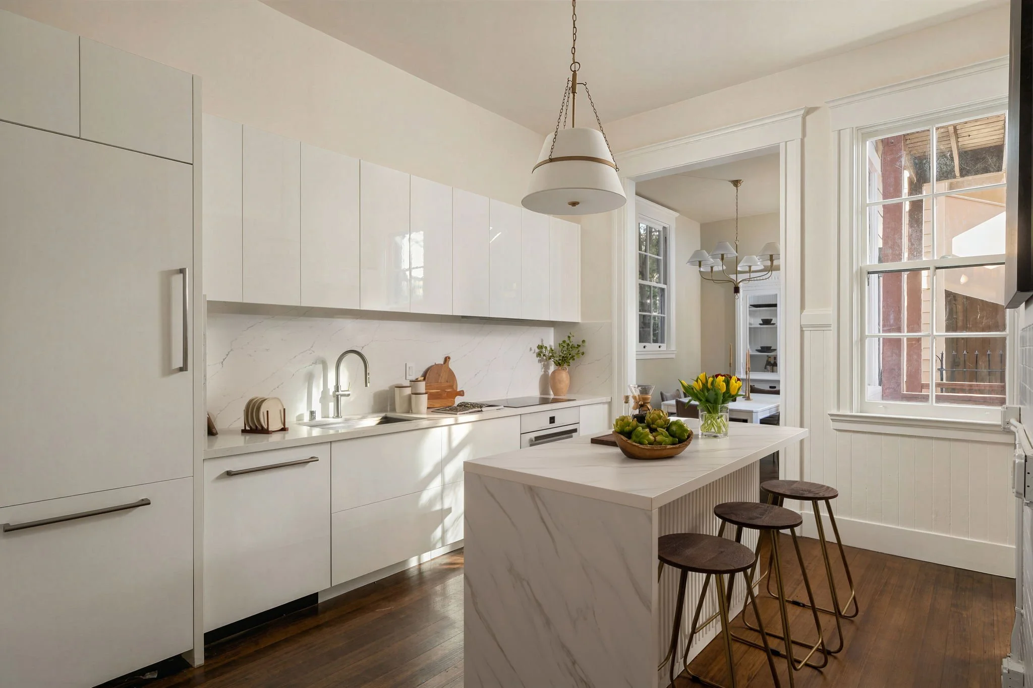 Modern white kitchen with marble countertops, pendant and chandelier lighting, large windows, a vase with flowers, and bar stools around a marble island.