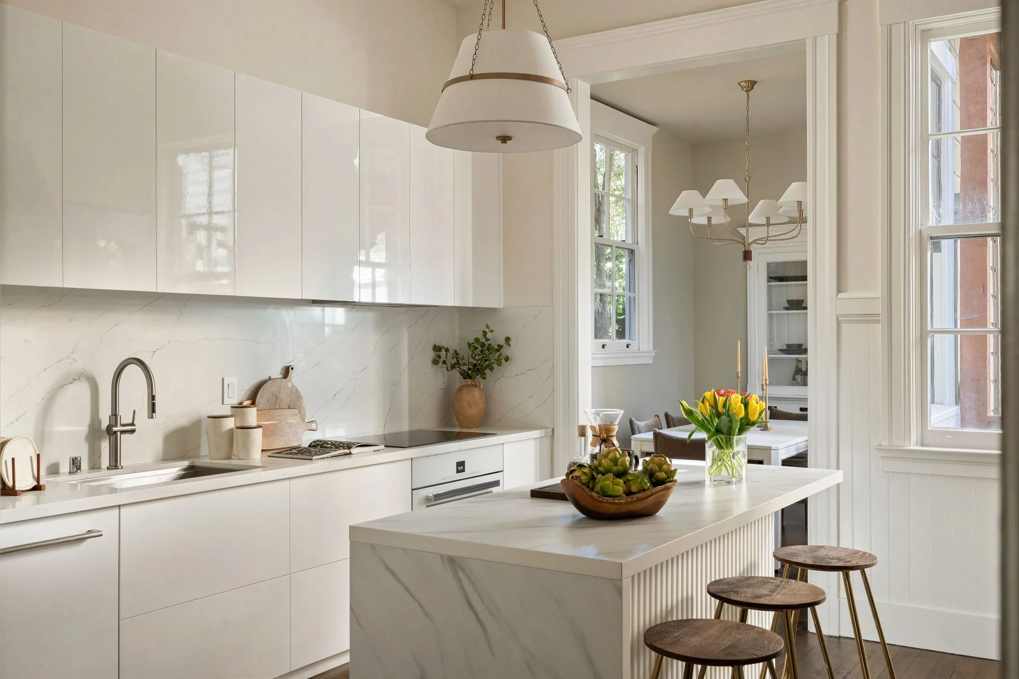 Modern kitchen with white cabinets and marble countertops, a table with a flower vase and artichokes, and wooden bar stools.