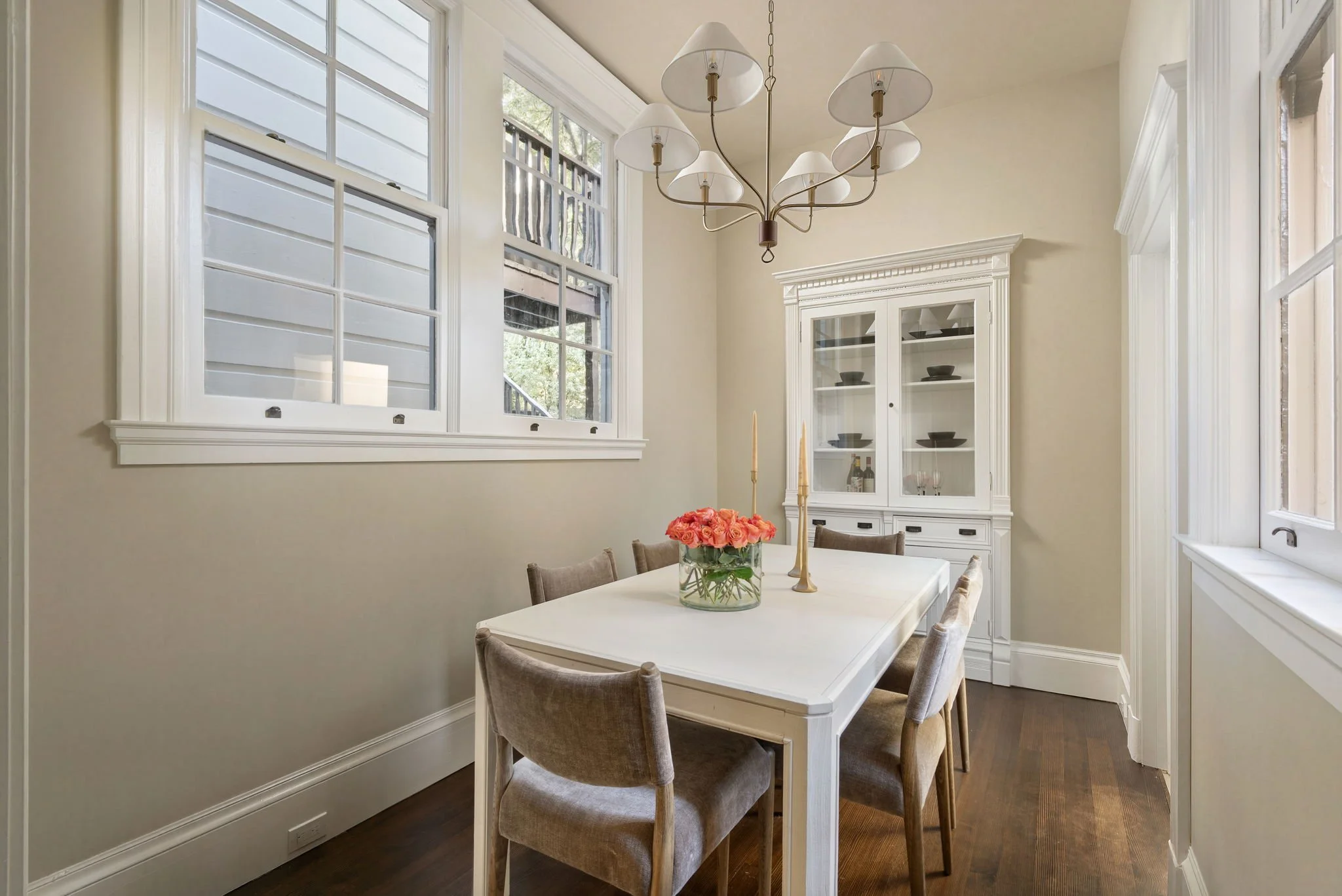 A dining room with a white table, beige upholstered chairs, a vase with pink roses, a white china cabinet, multiple windows, and a chandelier.