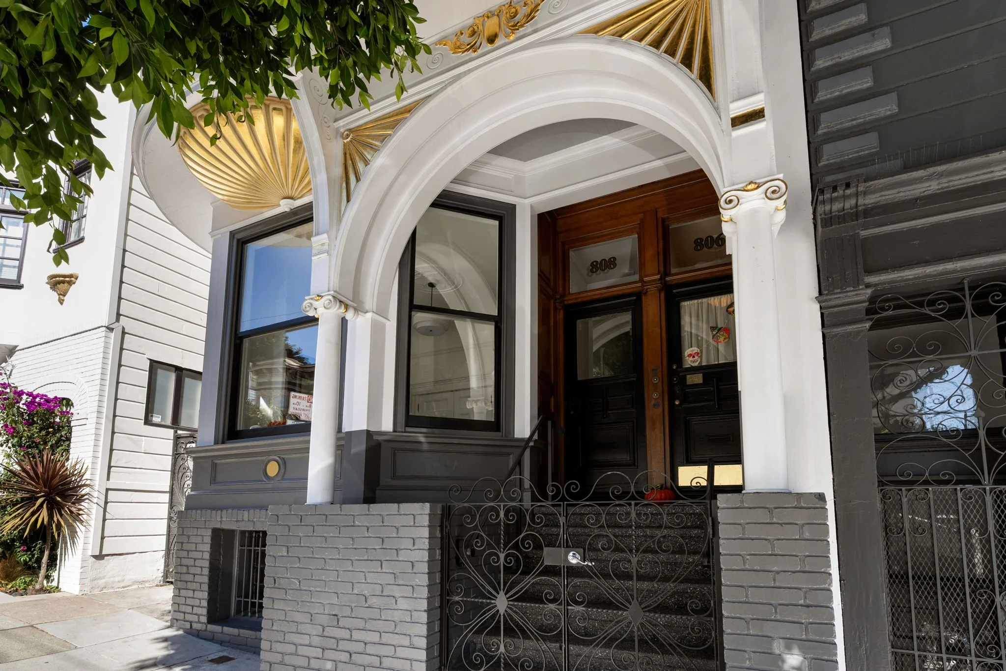Front entrance of a townhouse with white and brick exterior, arched doorway, wooden double doors, ornamental gold accents, black wrought iron gate, and decorative elements, with a tree and plants nearby.