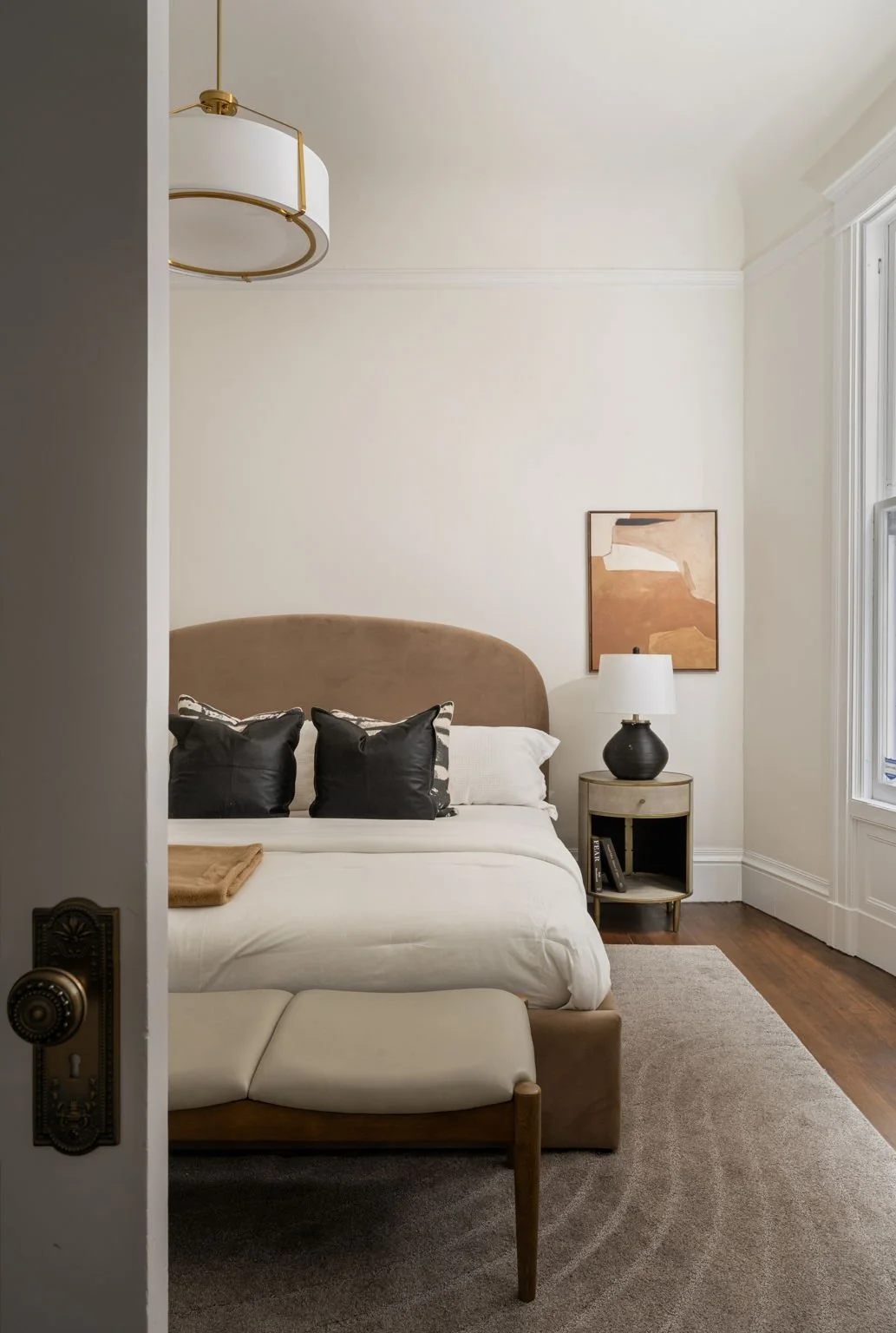View of a bedroom with a bed, black and white pillows, bedside table with a lamp, window, framed artwork, and a bench at the foot of the bed.