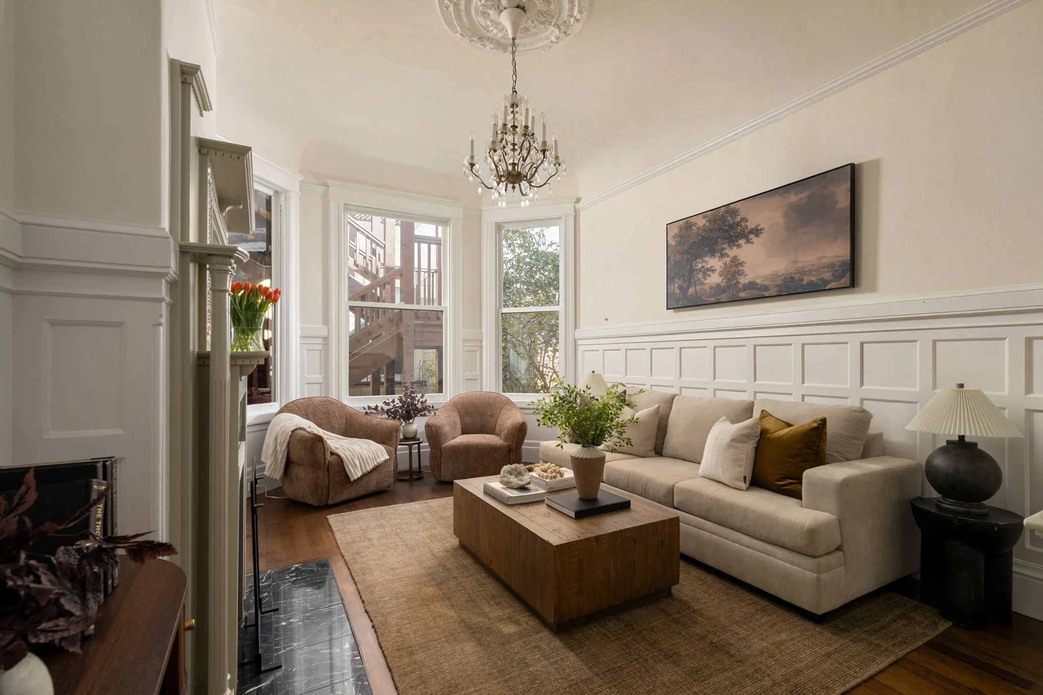Living room with beige sofa, pink armchairs, wooden coffee table, artwork of trees on the wall, chandelier, large windows, and decorative plants.