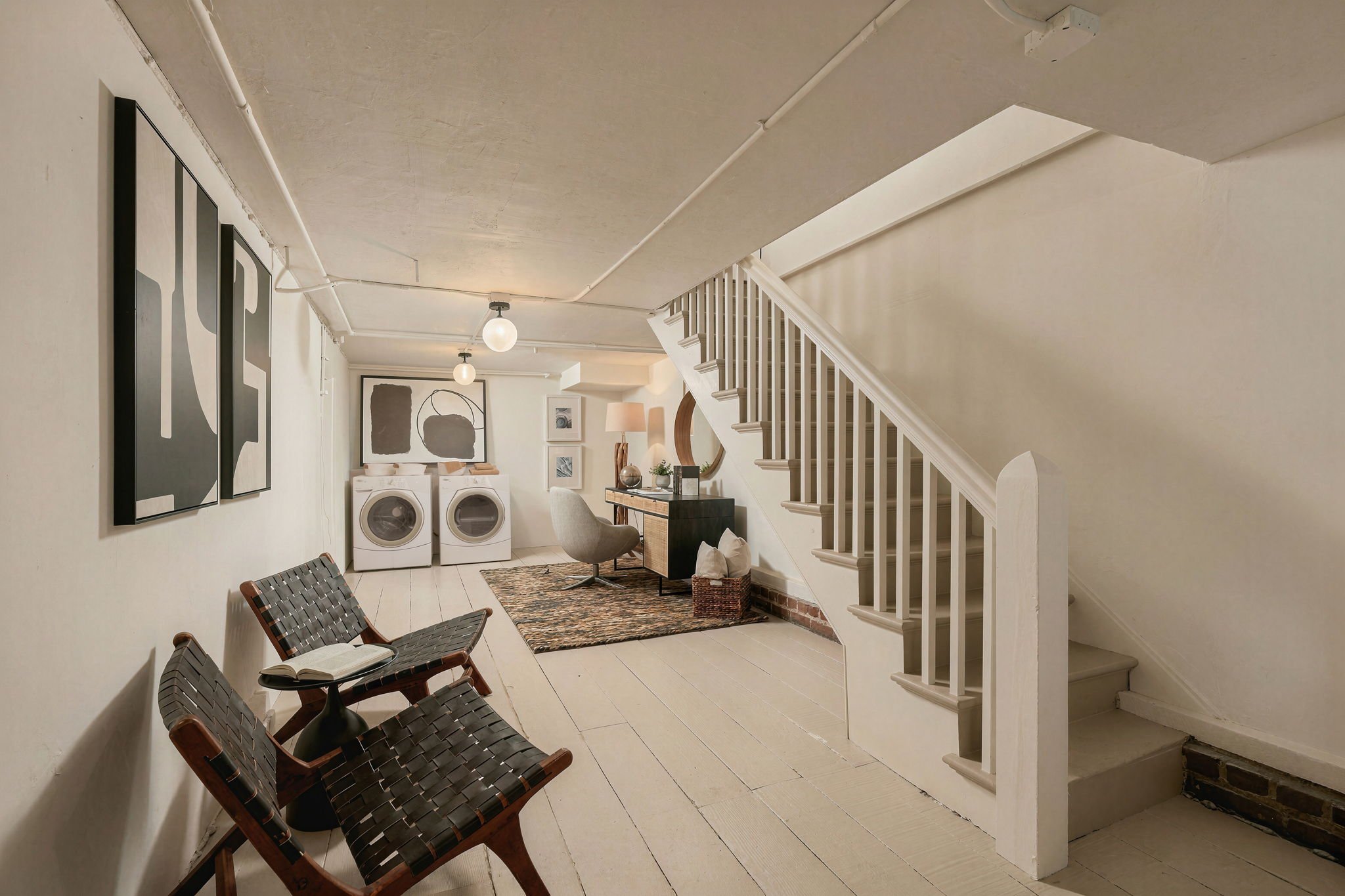 A laundry room with a white washer and dryer, black and tan chairs, artwork on the walls, and a staircase.
