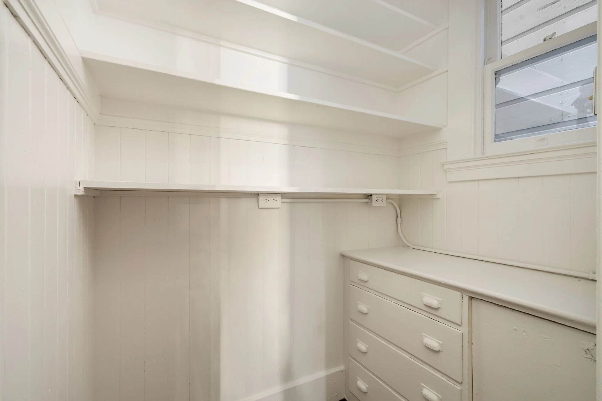 An empty white walk-in closet with shelves, drawers, and a window on the right wall.