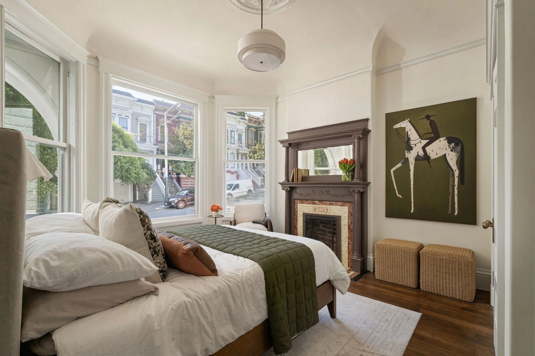 A cozy bedroom with a bed covered in white and green bedding, a large window showing a street with Victorian-style houses, a fireplace with a mirror above it, a vase with red tulips, and a painting of a black and white horse with a rider.
