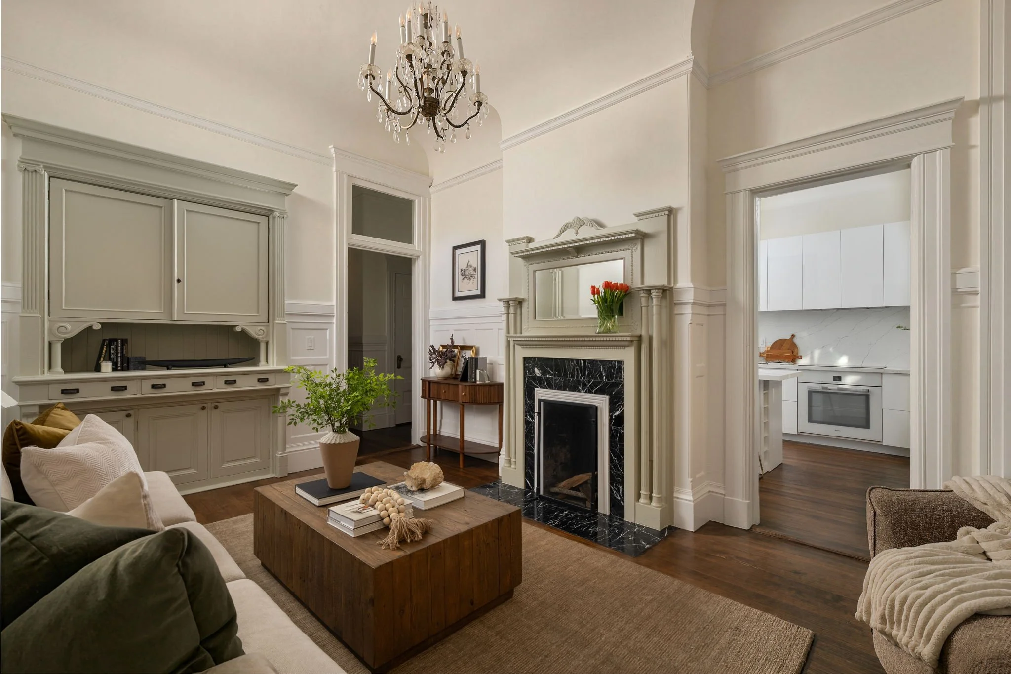Living room with fireplace, large mirror, and chandelier, with beige walls and white trim, decorated with plants, books, and neutral-colored furniture.
