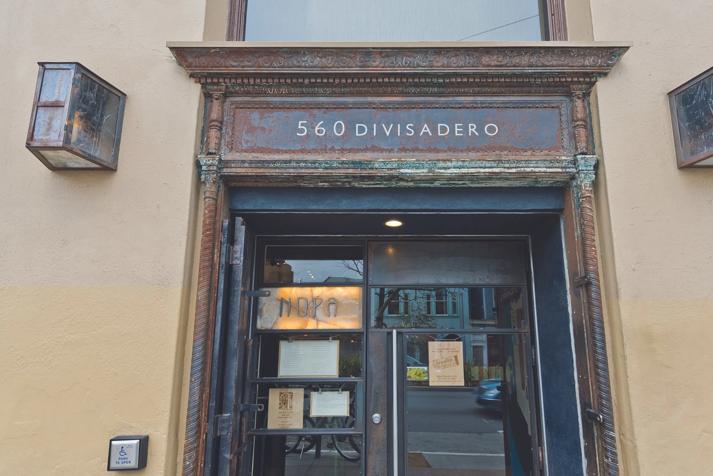 The image shows the front entrance of a building with a sign that reads "560 DIVISADERO." The entrance has a black-framed glass door with multiple notices and signs posted on it. There are two vintage-style wall-mounted light fixtures on either side 