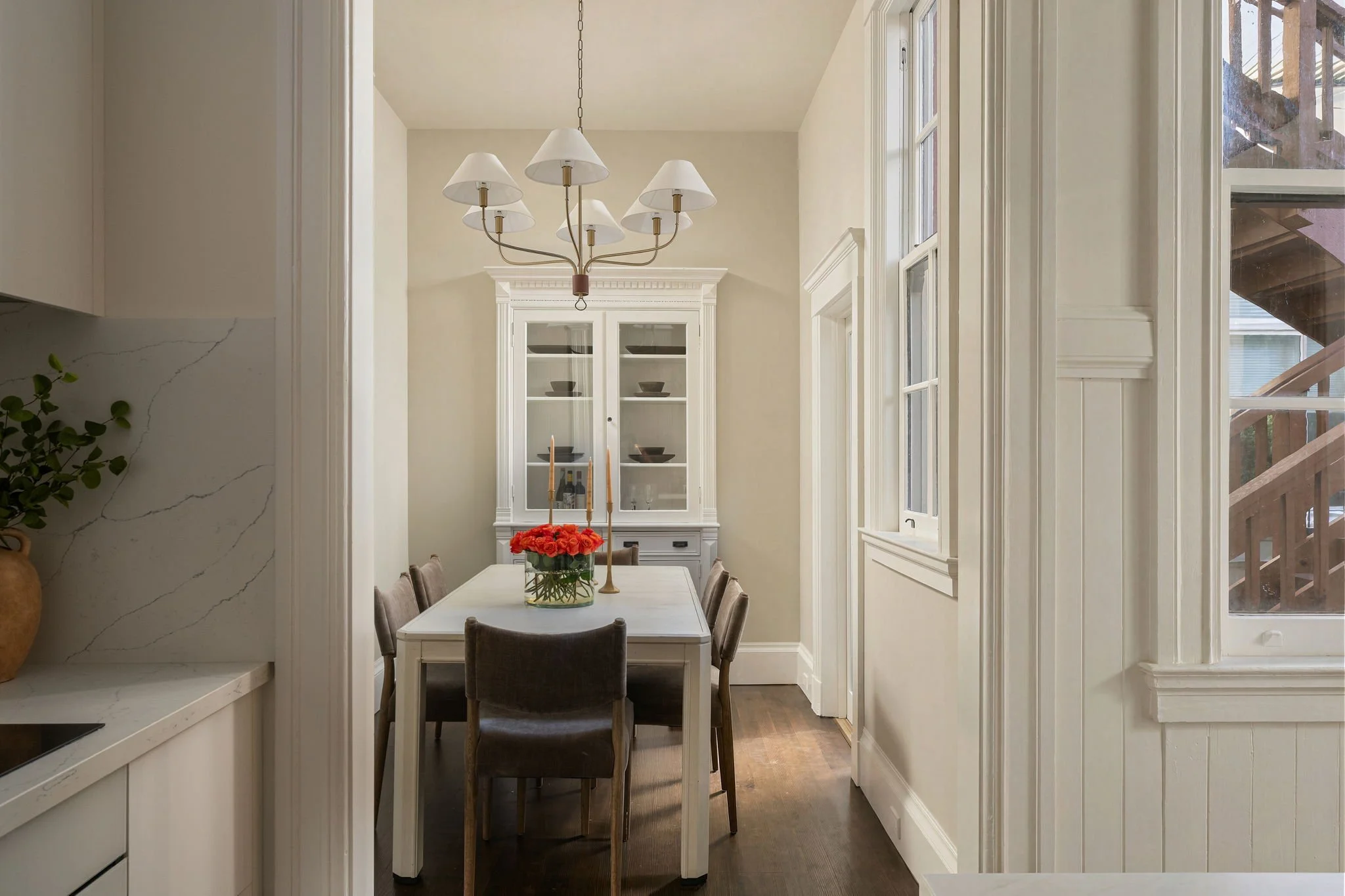 A dining room with a white dining table surrounded by six upholstered chairs, a flower arrangement, two candlesticks, a white china cabinet, and a chandelier hanging from the ceiling. Large windows with white trim let in natural light, and there is a staircase visible through the window.