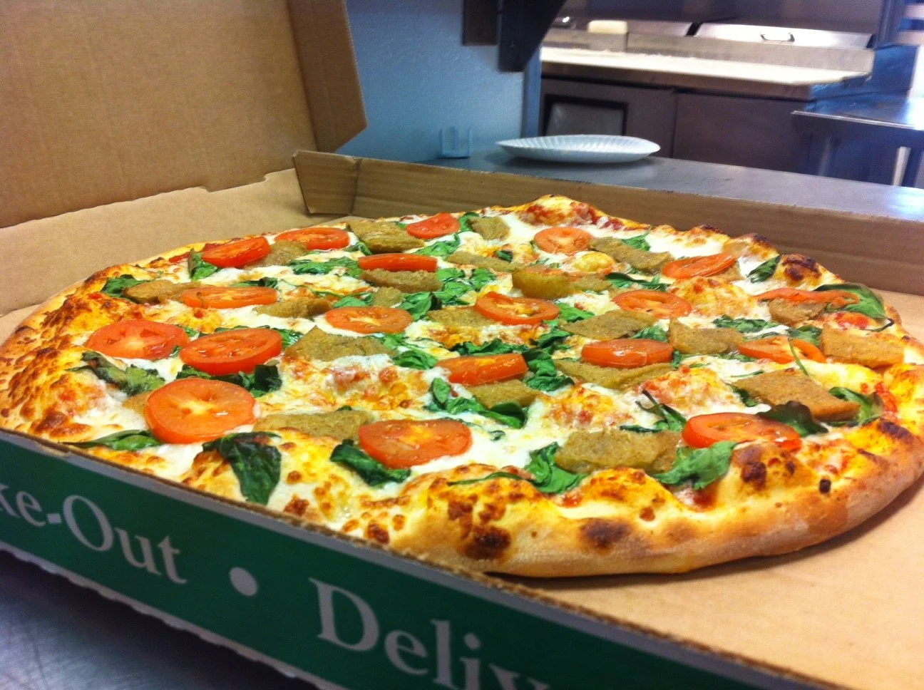 A fresh pizza in a cardboard box, topped with tomatoes, spinach, and breaded chicken pieces.