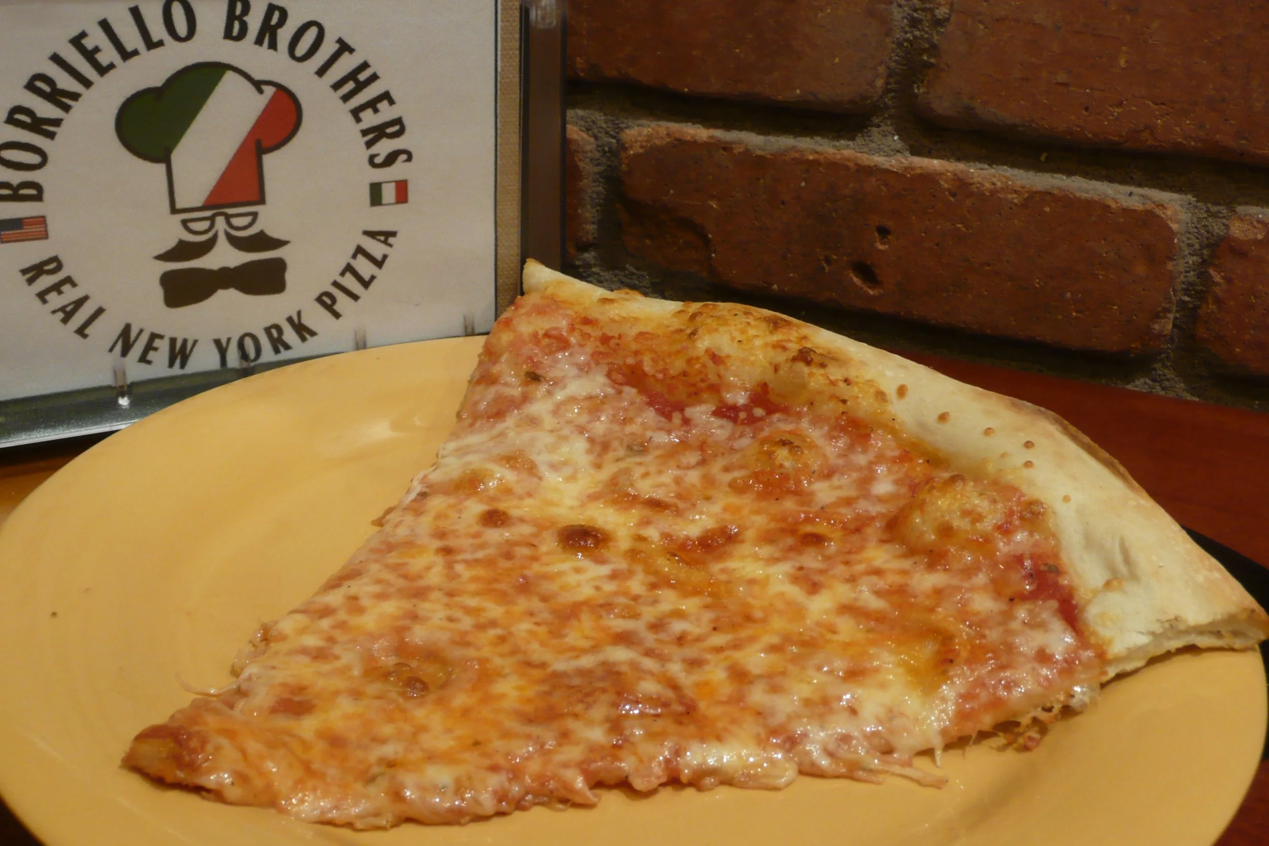 A slice of cheese pizza on a yellow plate in front of a brick wall, with a pizza place sign in the background that reads 'Borrieello Brothers Real New York Pizza.'