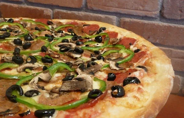 Close-up of a pizza topped with green bell peppers, black olives, mushrooms, and slices of pepperoni, placed against a brick wall background.