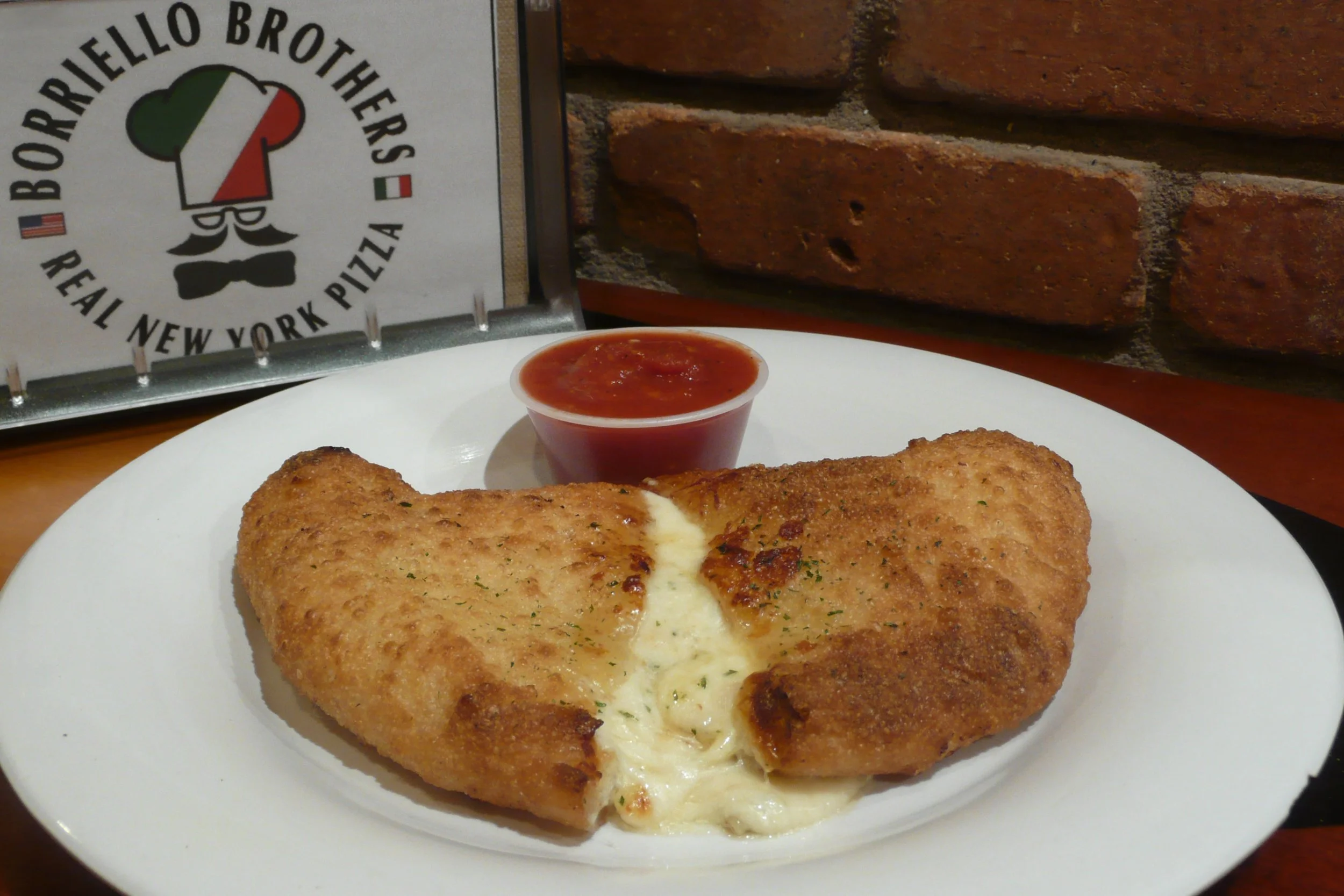 Fried mozzarella stick on a white plate with melted cheese inside, accompanied by a small cup of tomato sauce. In the background, a sign with a chef's hat logo and the words 'Borrello Brothers Real New York Pizza' is visible, as well as a brick wall.