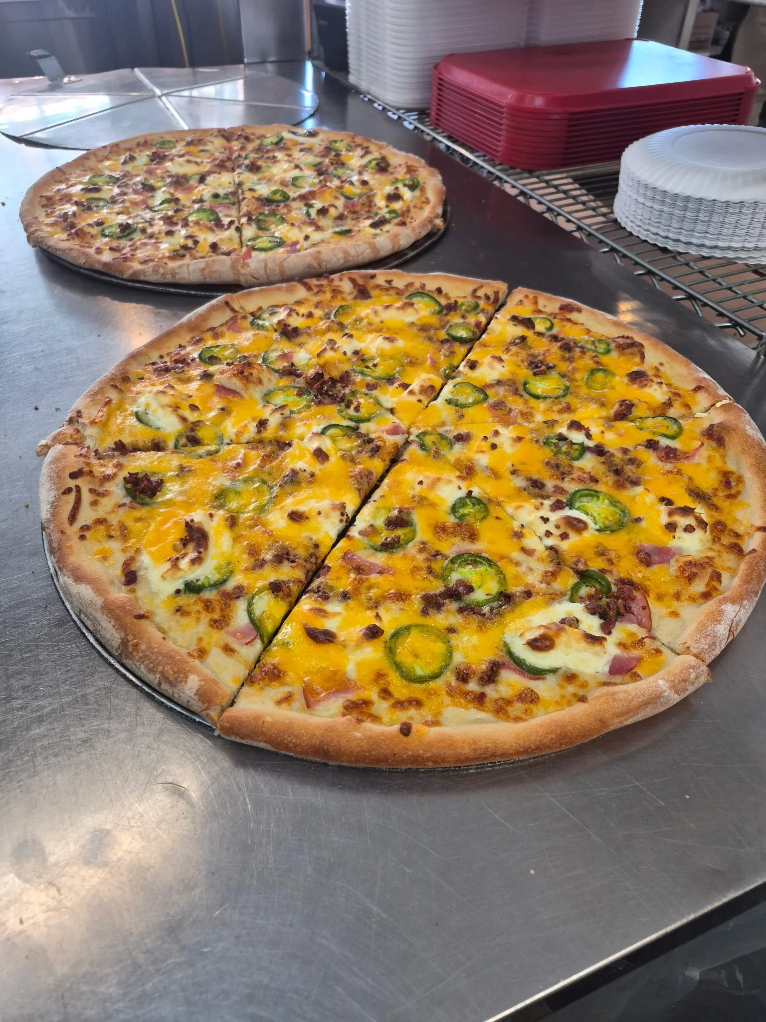 Two freshly baked pizzas on a metal countertop, one with cheese, jalapeños, and bacon, and the other with cheese, bacon, mushrooms, and green peppers, in a restaurant kitchen setting.
