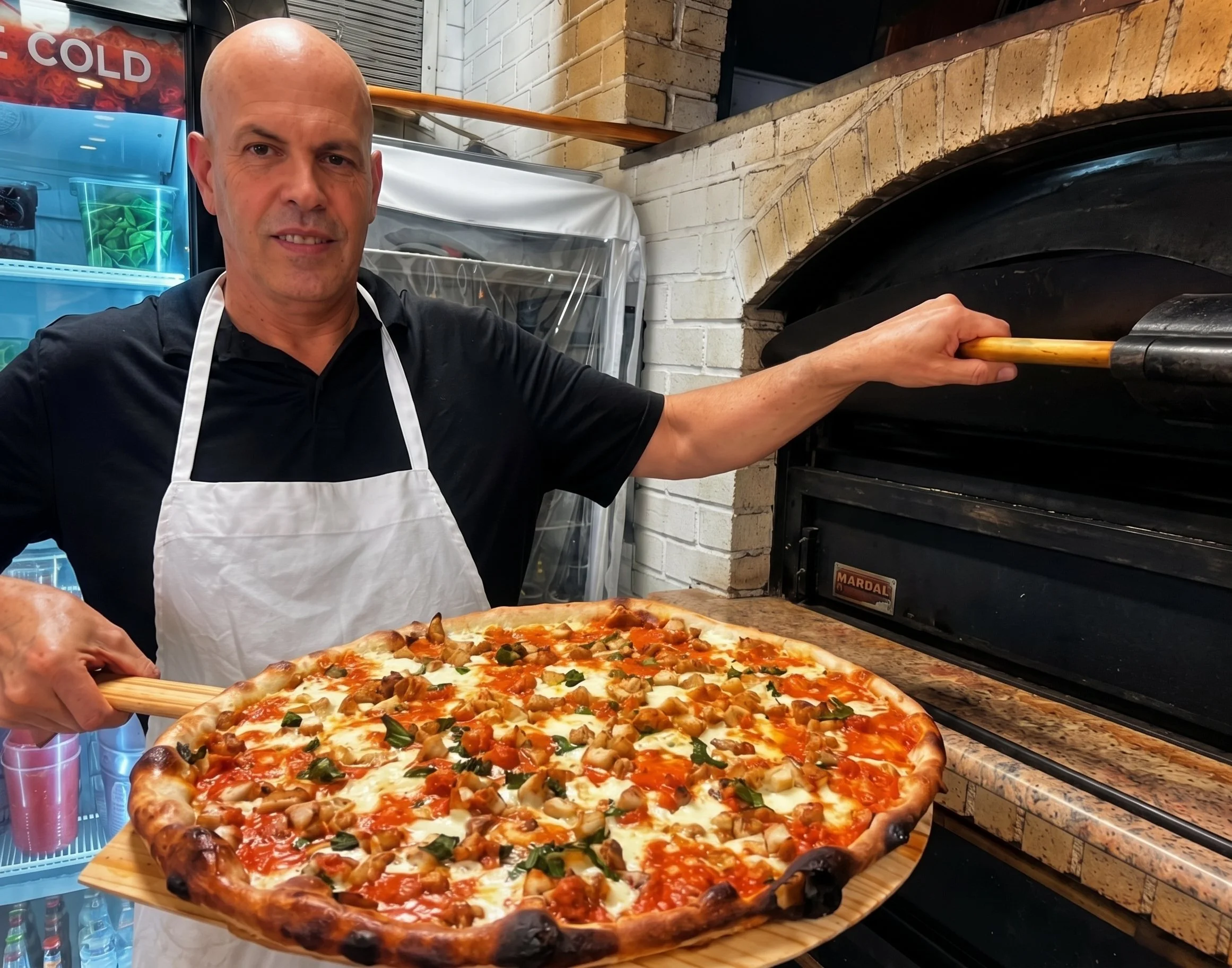 A man with a bald head wearing a black shirt and white apron is taking a large pizza out of an oven. The pizza has cheese, chicken, and vegetables toppings.