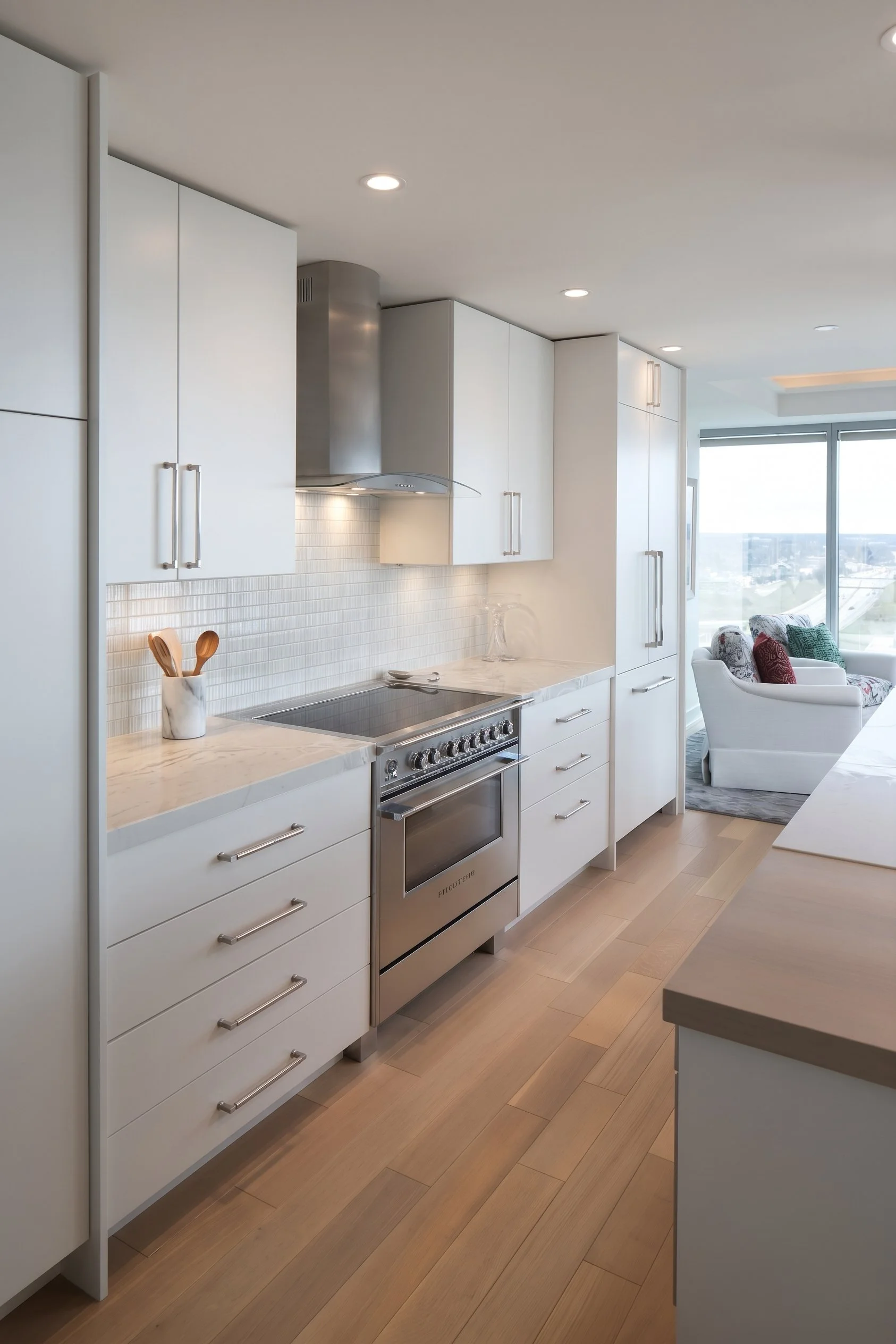 Modern white kitchen with stainless steel range, marble countertops, tile backsplash, and floor-to-ceiling windows overlooking the city