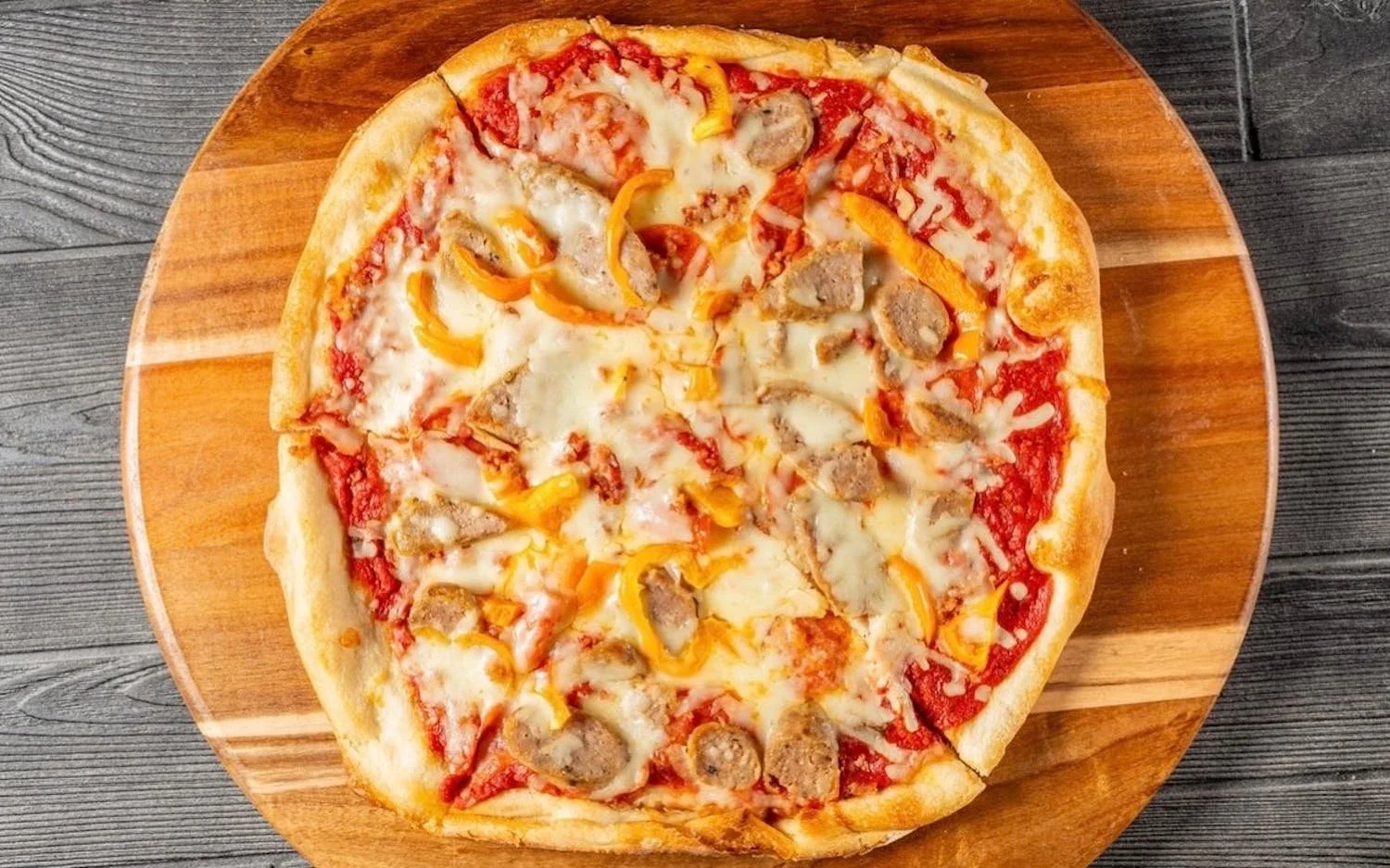Pizza with sausage, yellow bell peppers, and cheese on a wooden serving board.
