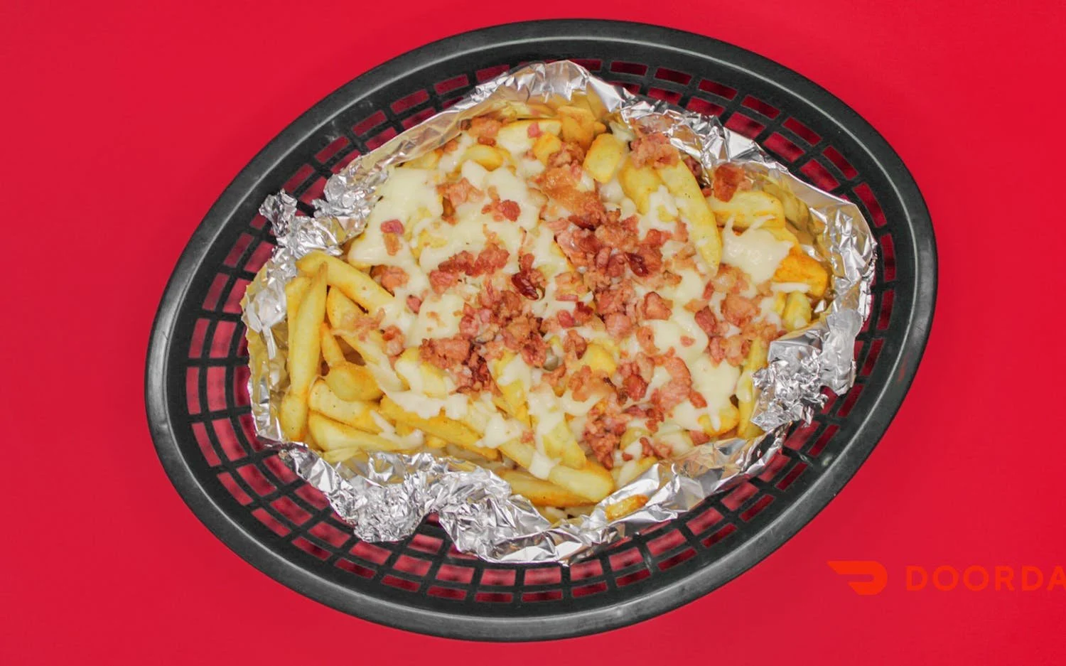 French fries topped with shredded cheese and bacon bits in a black basket lined with aluminum foil, on a red background.