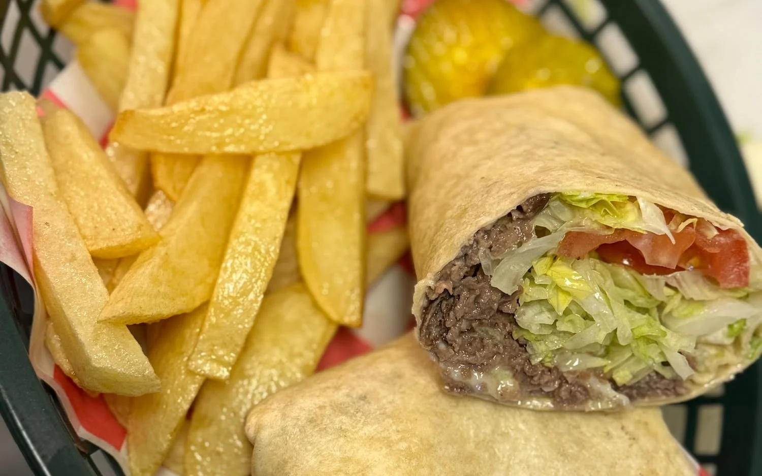 A cheeseburger wrap with lettuce, tomato, and ground beef, cut in half, served with a side of French fries in a basket.