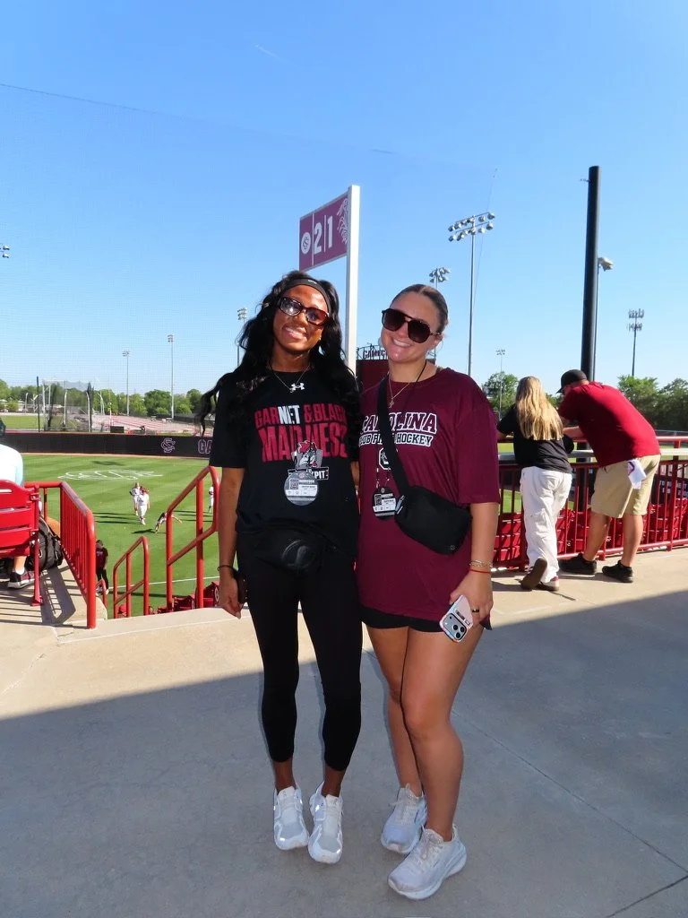 Two women standing together at a sports stadium, smiling. One wears a black T-shirt and leggings, the other a maroon T-shirt and shorts, both with sunglasses and sneakers. The background shows a football field and blue sky.
