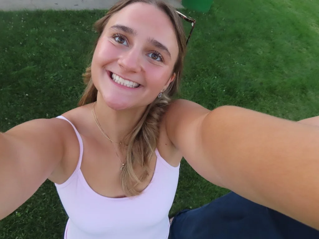 A young woman with long hair and a white tank top taking a selfie outdoors on a grassy area, smiling broadly.