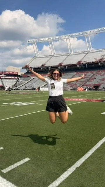 A woman jumping in the air on a football field at a stadium, with arms outstretched and a big smile, under a partly cloudy sky.