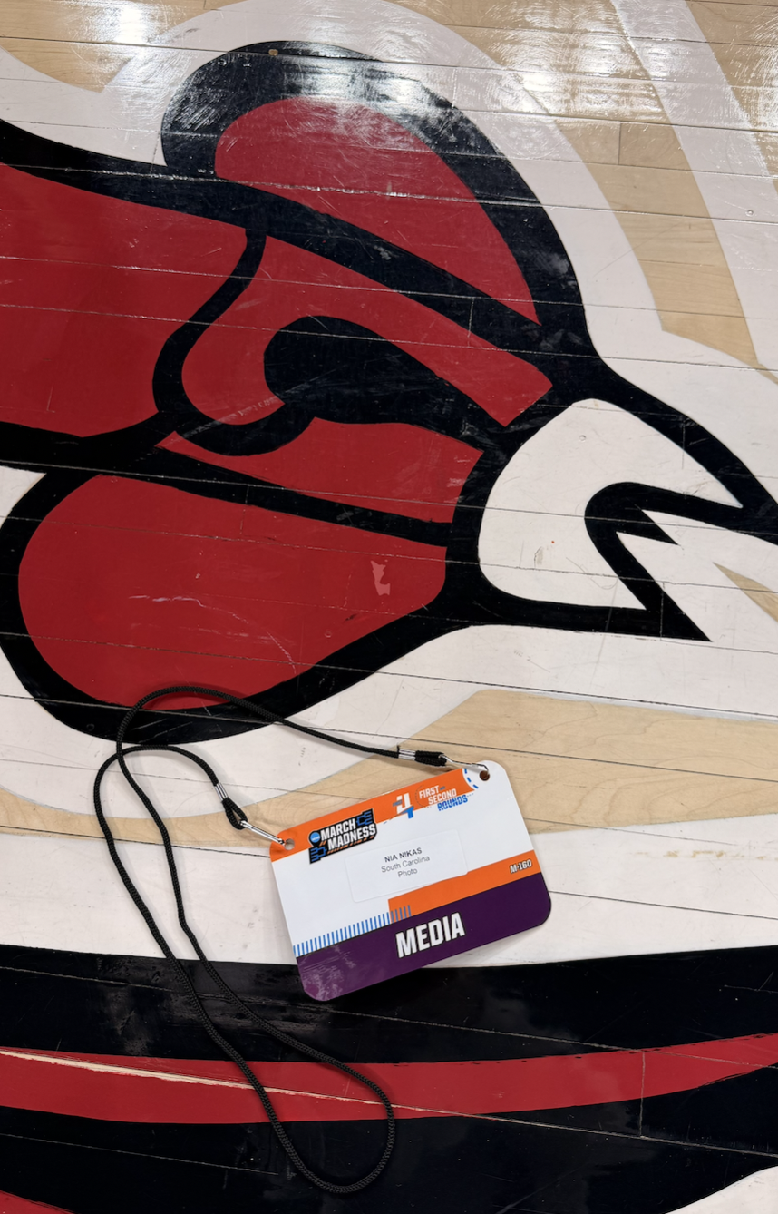 A basketball court floor with a large painted logo of a red bird wearing a black hat and red clothes. There is a media credential badge with a black lanyard on the floor, labeled "Media" for the March Madness basketball tournament.