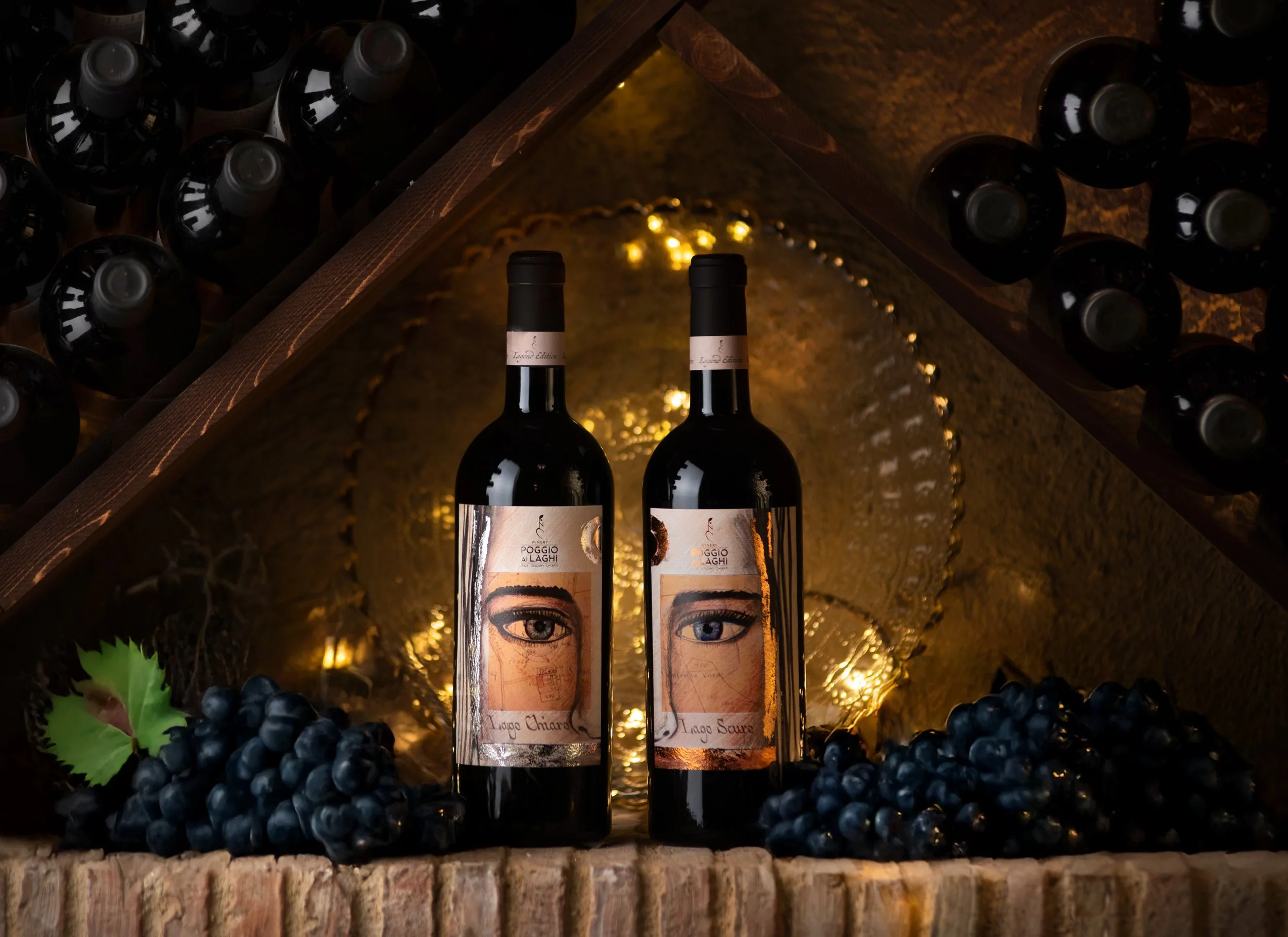 Two bottles of red wine are displayed on a reflective surface, with a bunch of black grapes and green grape leaves in front. The background features a mirror and warm lighting, creating a cozy ambiance.